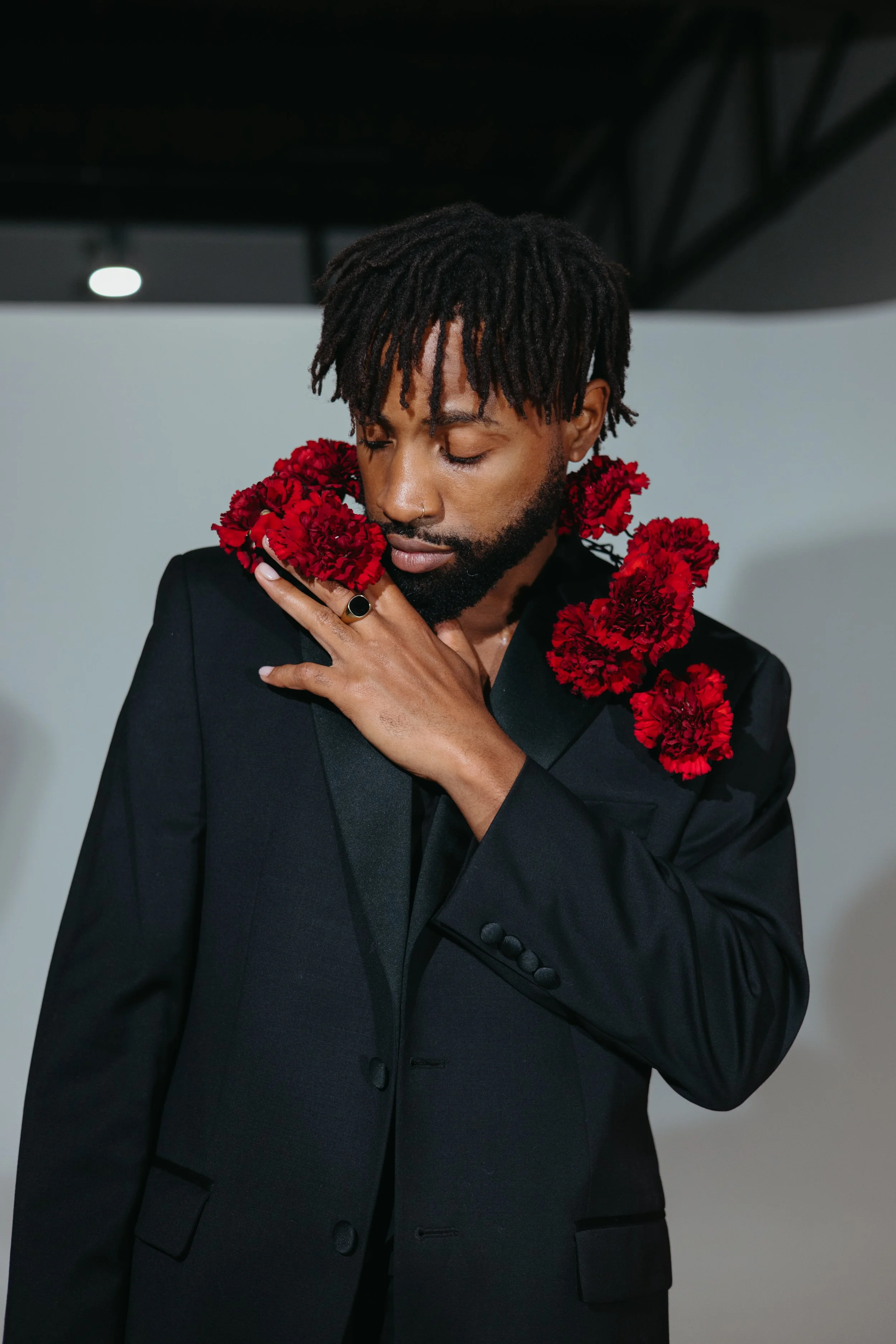 a Black male model in a dark suit against a background of white with a red backdrop and red carnations and roses
