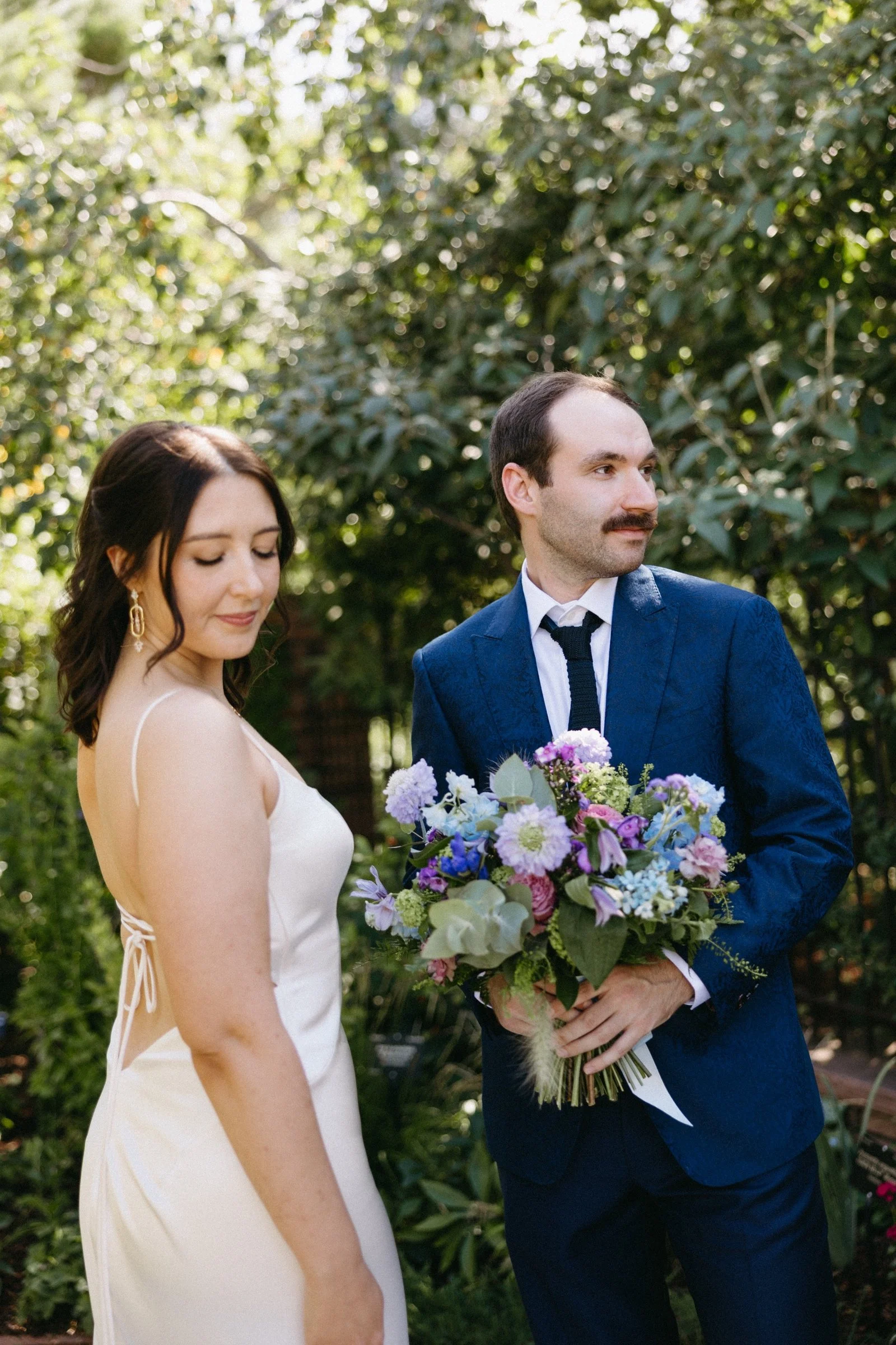 A woman in a white dress standing next to a man in a blue suit holding a colorful bouquet, outdoors with lush green trees in the background.