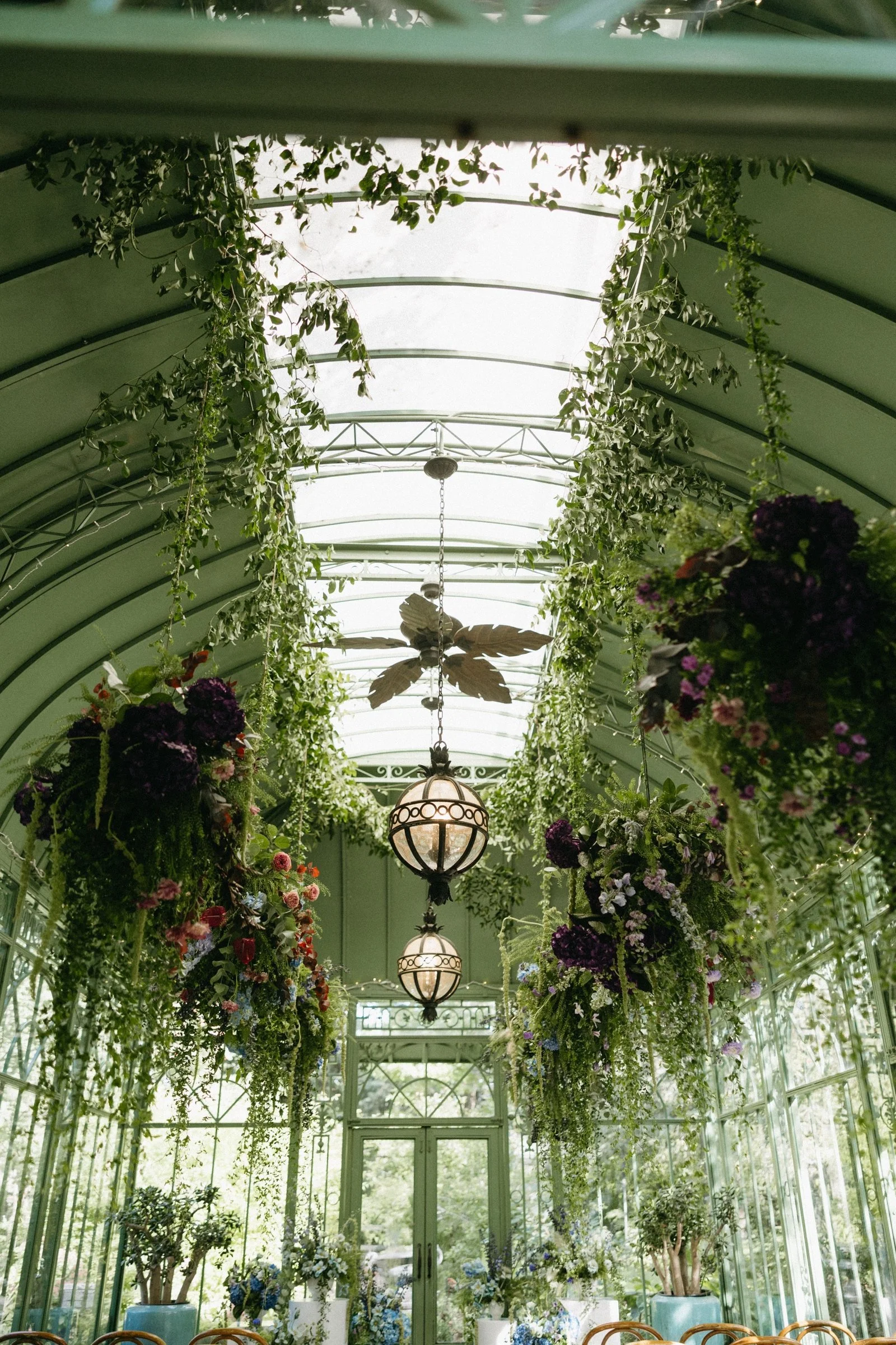 Interior of a glass conservatory decorated with hanging baskets of flowers and green foliage, vintage chandeliers, and ceiling fans.