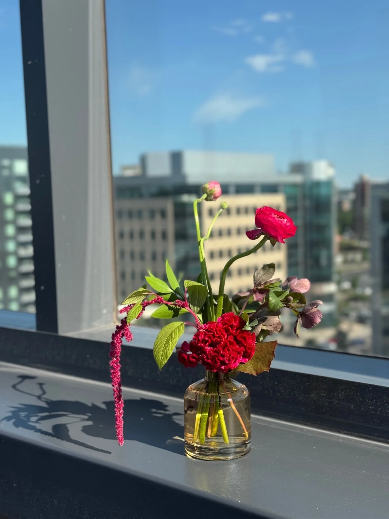 A small glass vase with a bouquet of red and pink flowers, including a pink tulip, a red cockscomb, and a pink astilbe, sitting on a windowsill with a cityscape in the background on a sunny day.