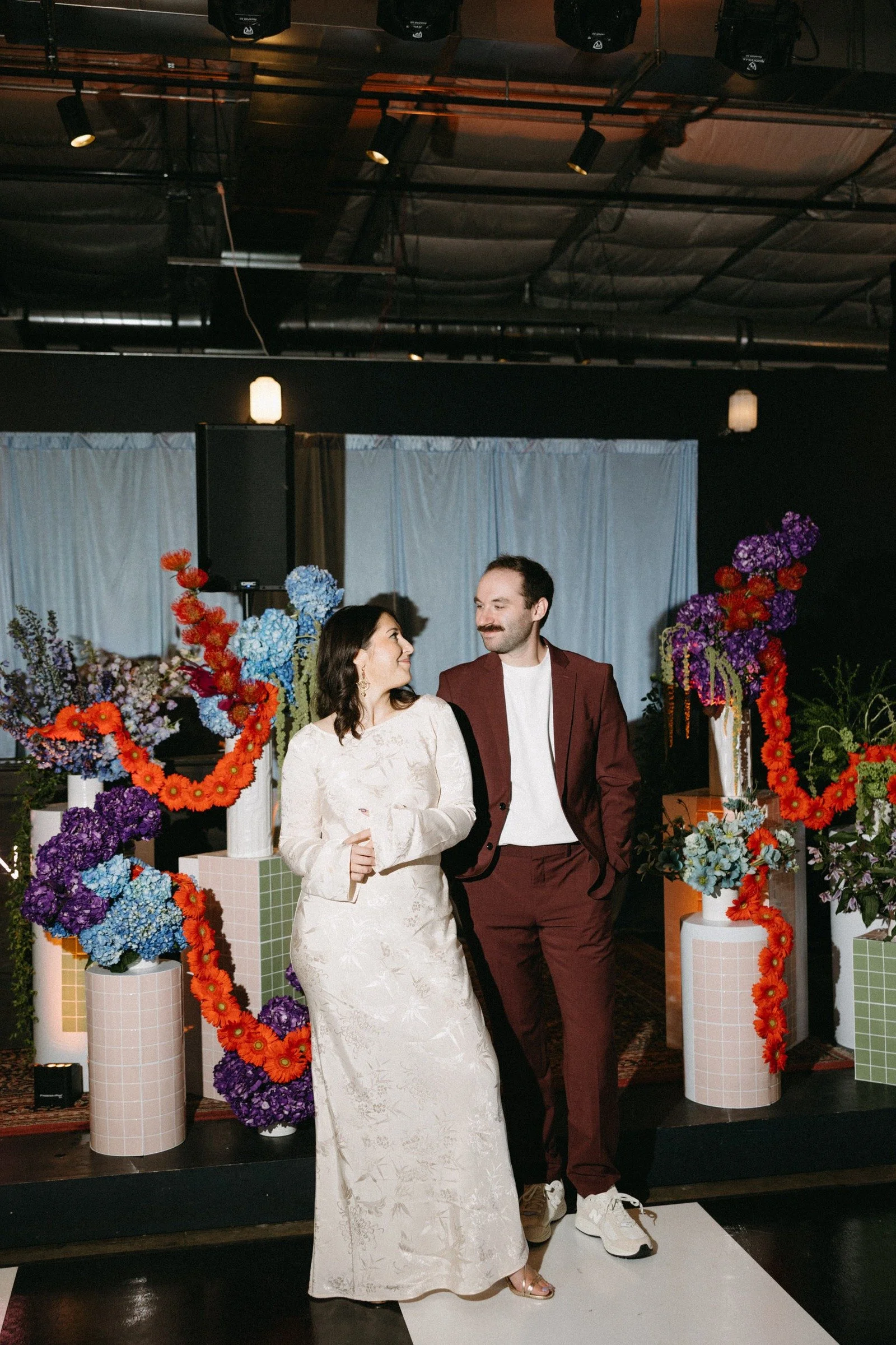 A man and woman in wedding attire standing in front of colorful flower arrangements and decorations. The woman wears a white dress and the man is dressed in a burgundy suit with a white shirt, several people at the wedding reception.