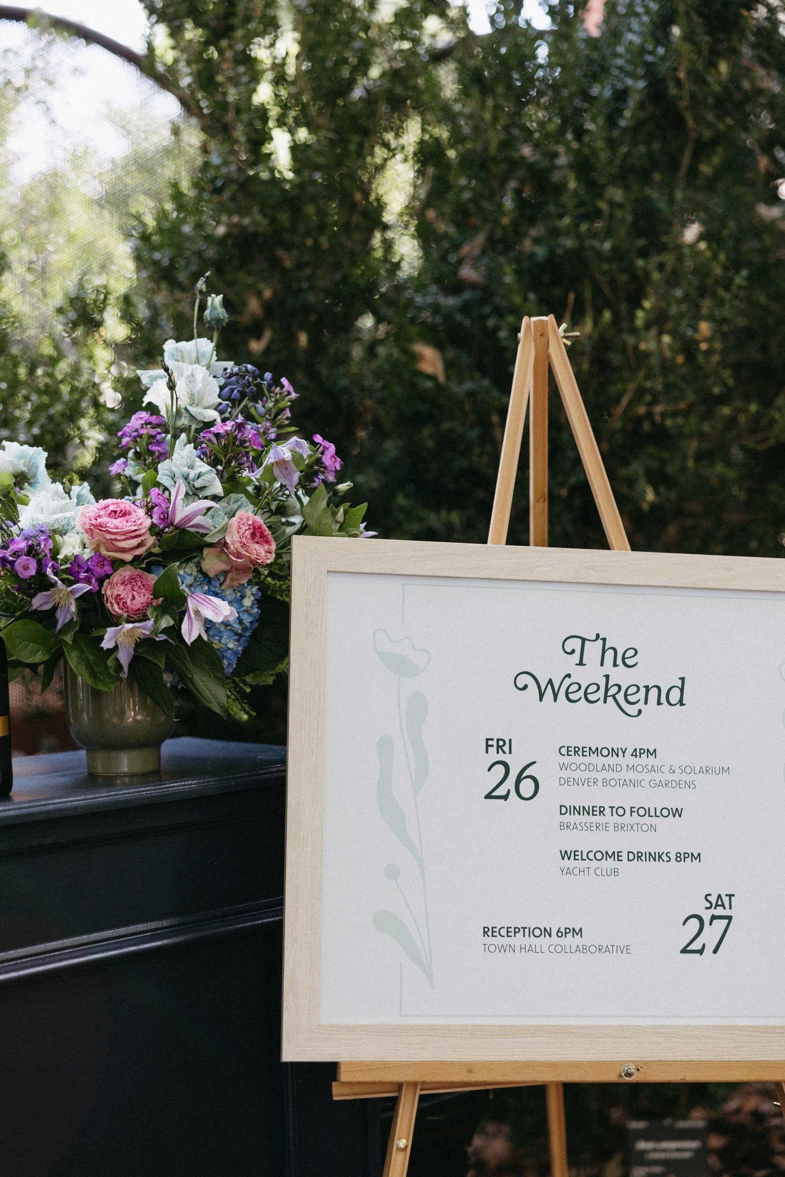 A white event schedule board on a wooden easel next to a flower arrangement in a grey vase at an outdoor event with green trees in the background.