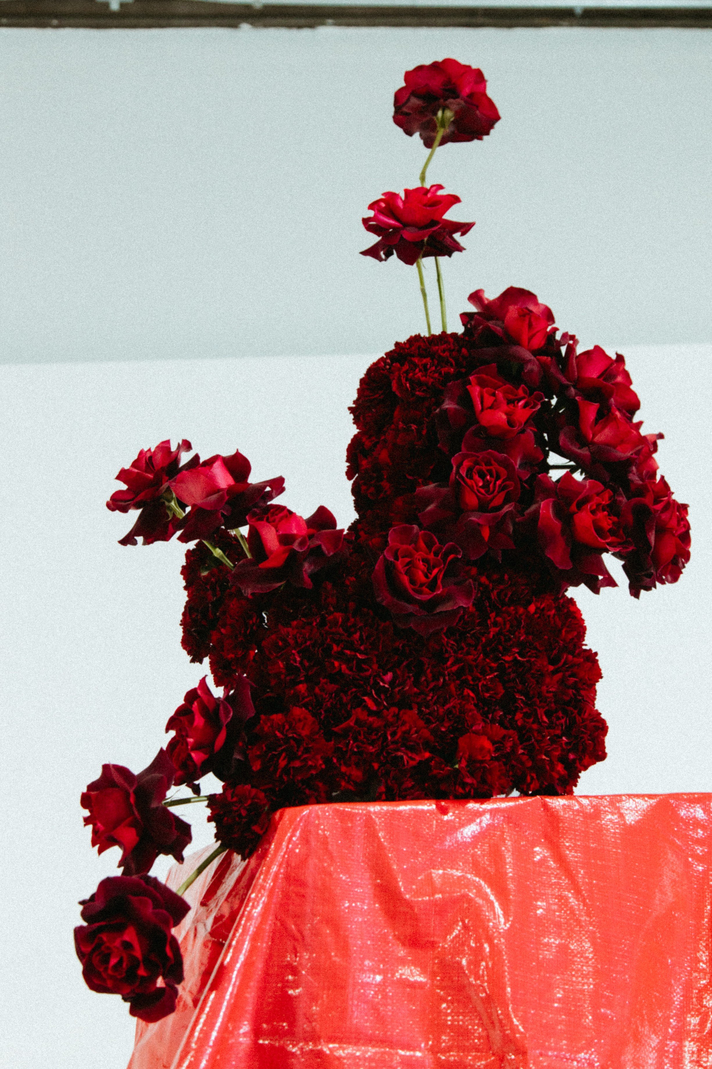 A flower arrangement with red roses and dark red carnations on a red, shiny, textured surface against a plain light background.