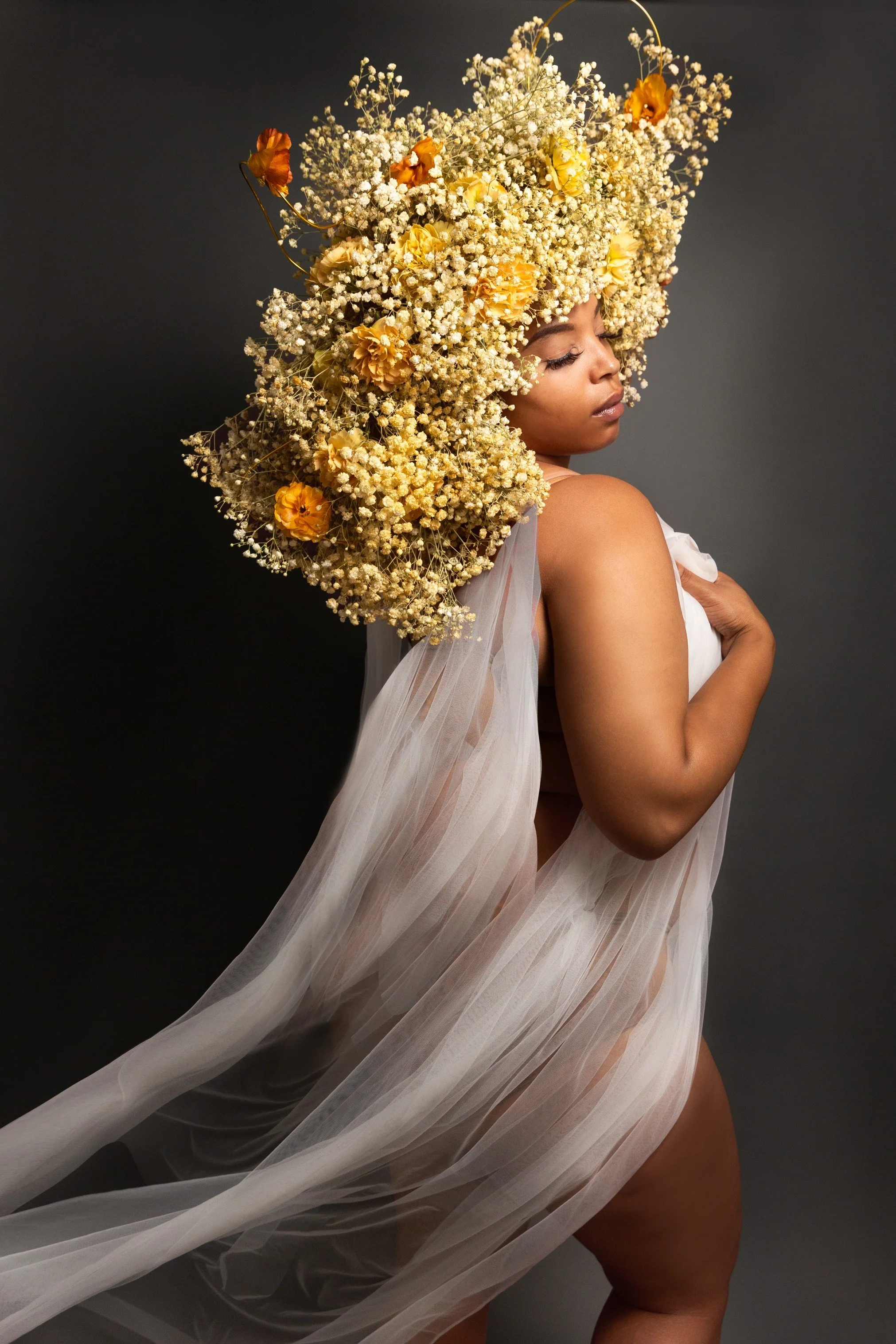 A woman with a large floral headpiece made of yellow and orange flowers and baby's breath, wearing a sheer, flowing fabric, standing against a dark background.
