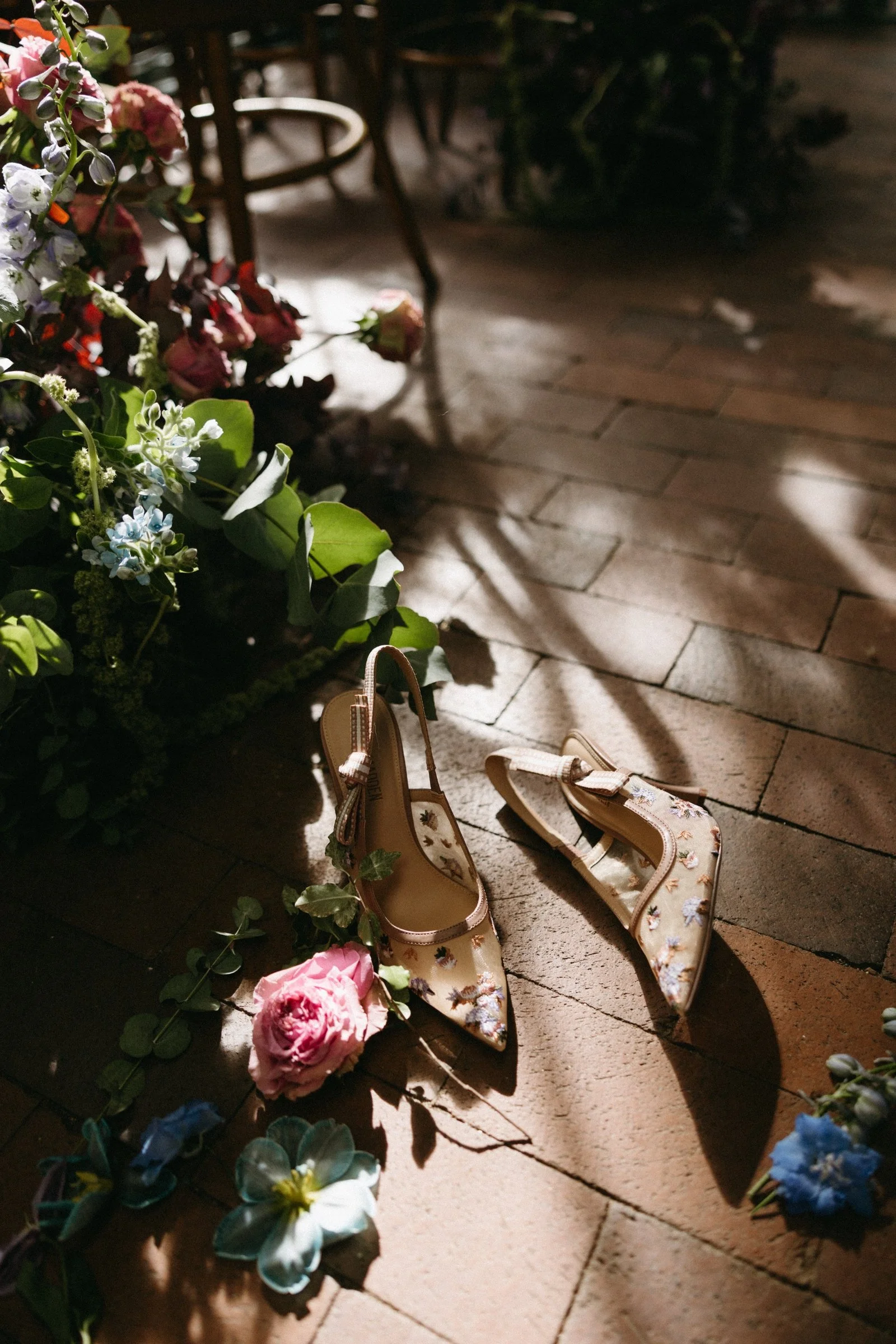 Pair of floral embroidered beige high heels shoes on a brick floor surrounded by pink, blue, and purple flowers and green foliage, with shadows cast across the scene.