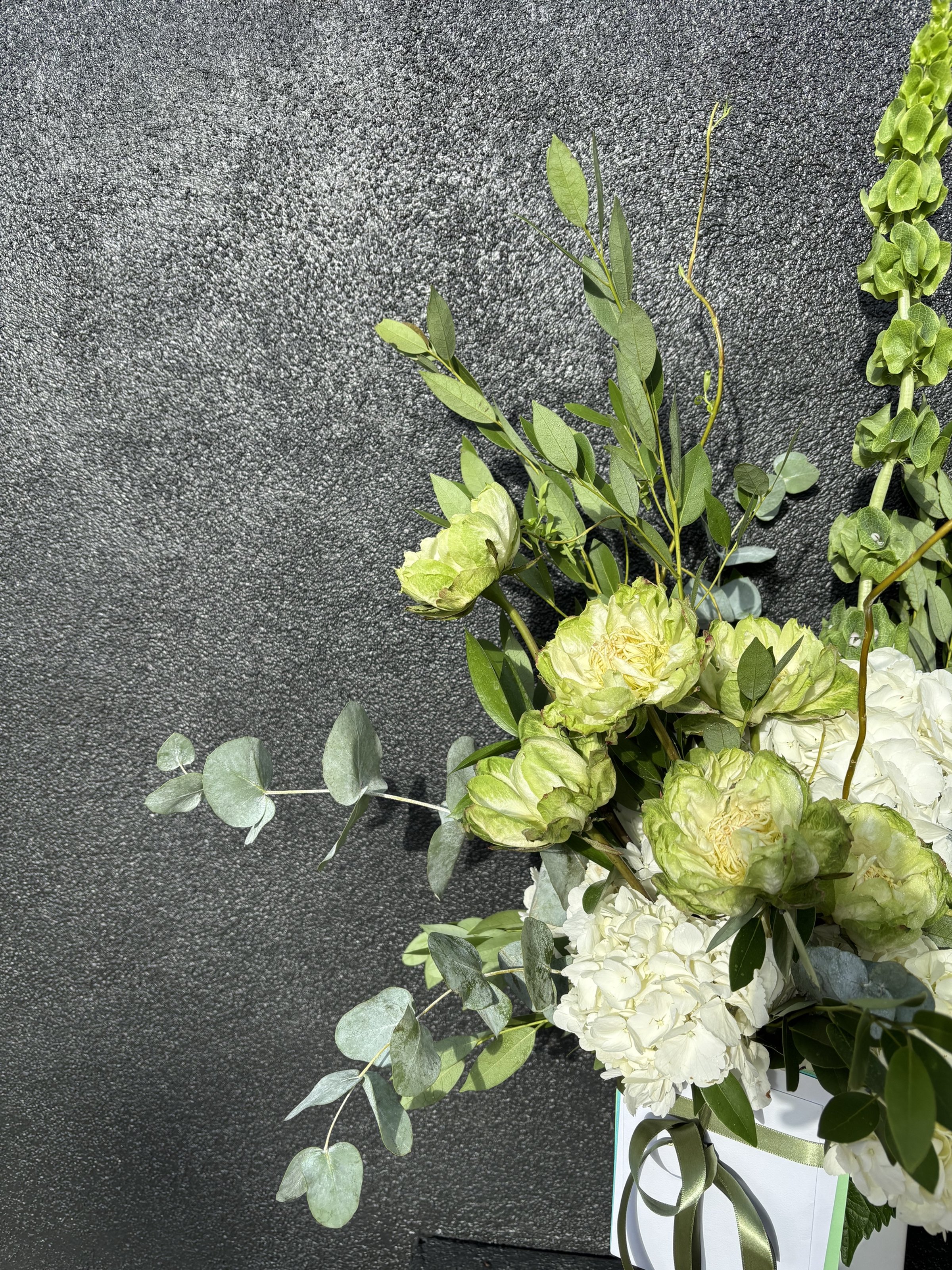 Arrangement of white and light green flowers with green foliage, tied with a green ribbon, against a textured dark background.