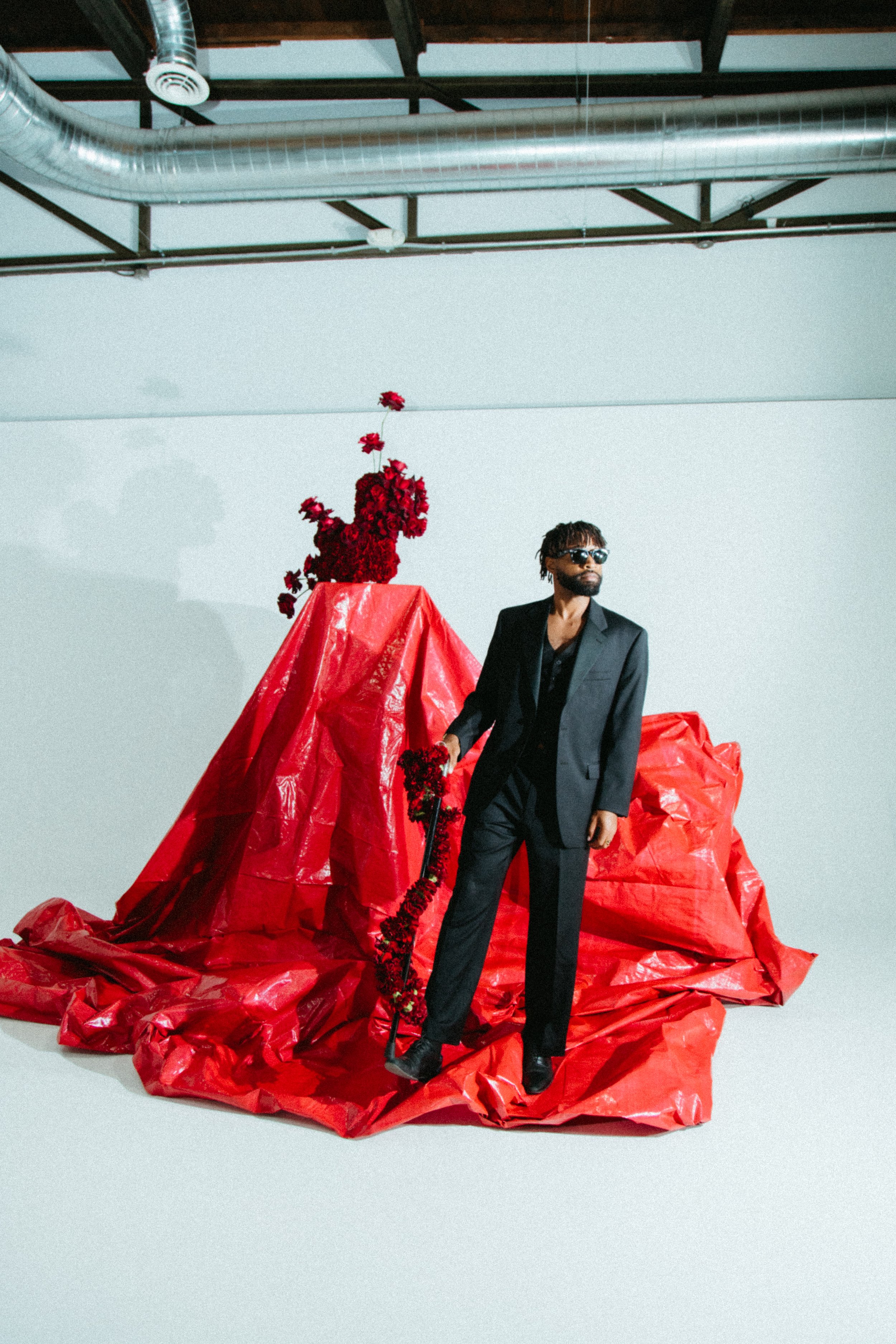 a Black male model in a dark suit against a background of white with a red backdrop and red carnations and roses