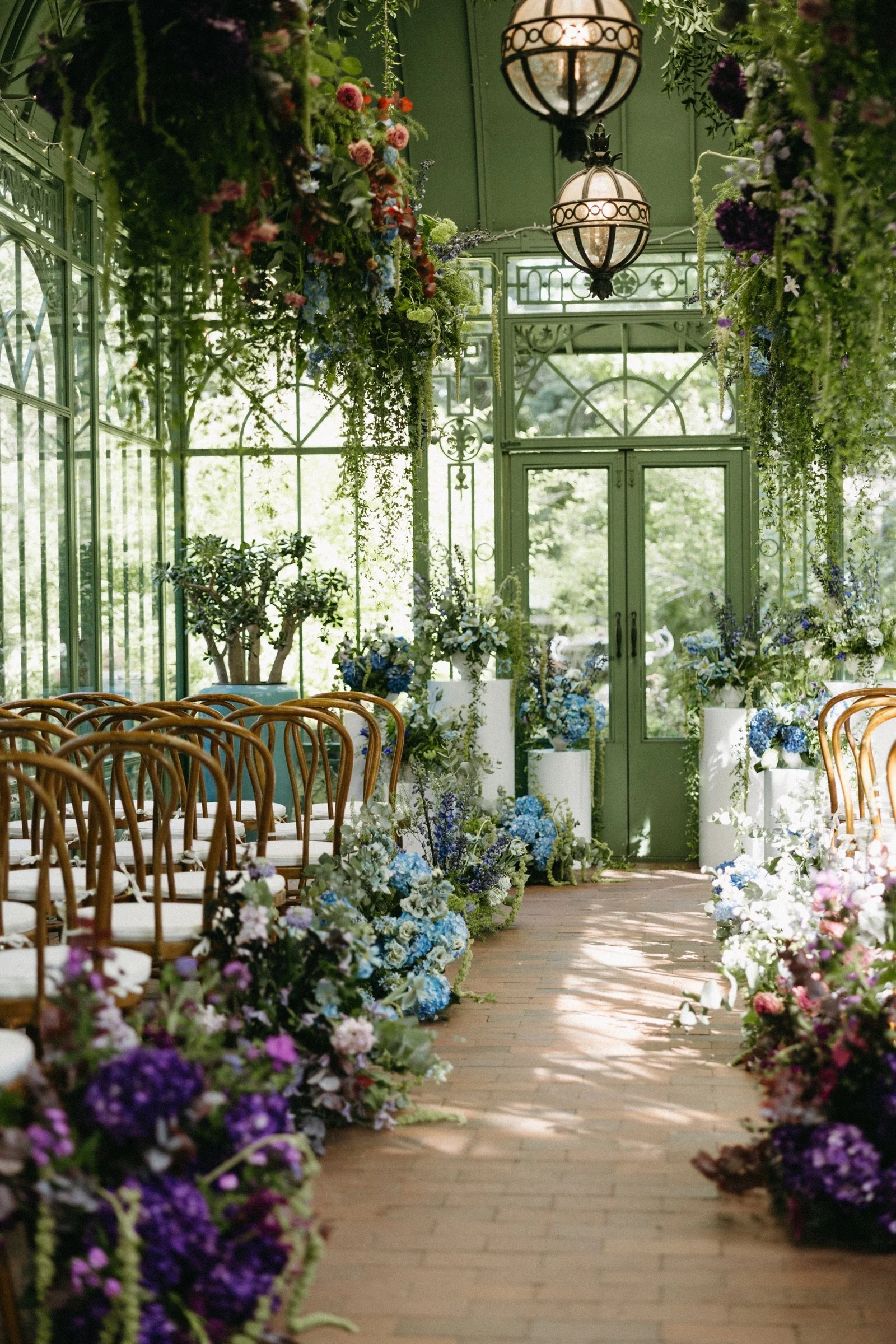Indoor wedding ceremony setup with green glass walls, wooden chairs with white cushions, and lush floral arrangements in shades of purple, blue, pink, and white, featuring hanging greenery and large vases, under ornate hanging lanterns.