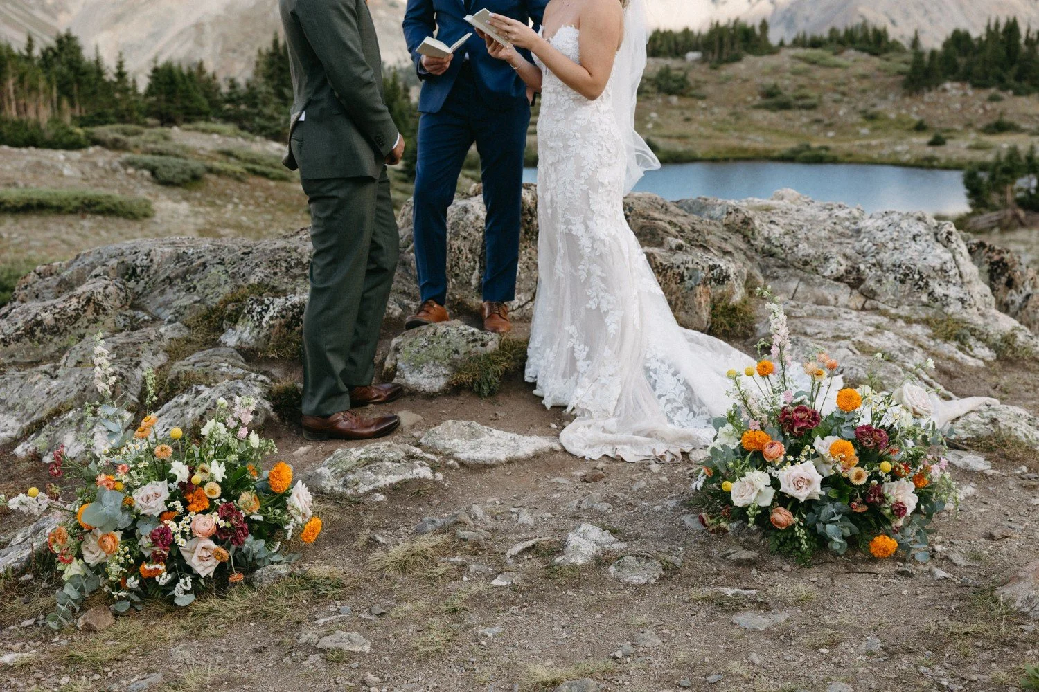 A couple getting married outdoors on a rocky landscape with two officiants. The bride is in a white lace wedding dress, and the groom is in a gray suit. Flowers are placed on the ground near them, and a lake and mountains are visible in the background.