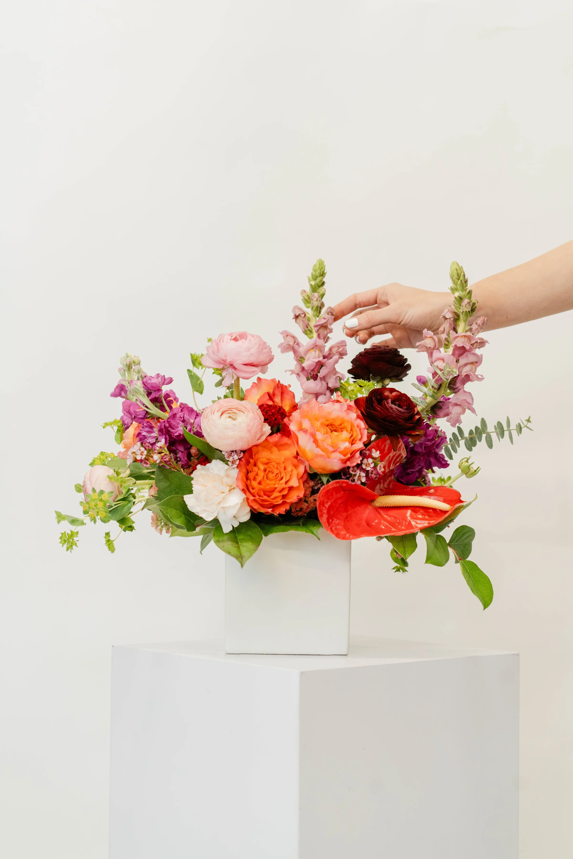 A hand arranging a colorful bouquet of flowers in a white square vase on a white pedestal