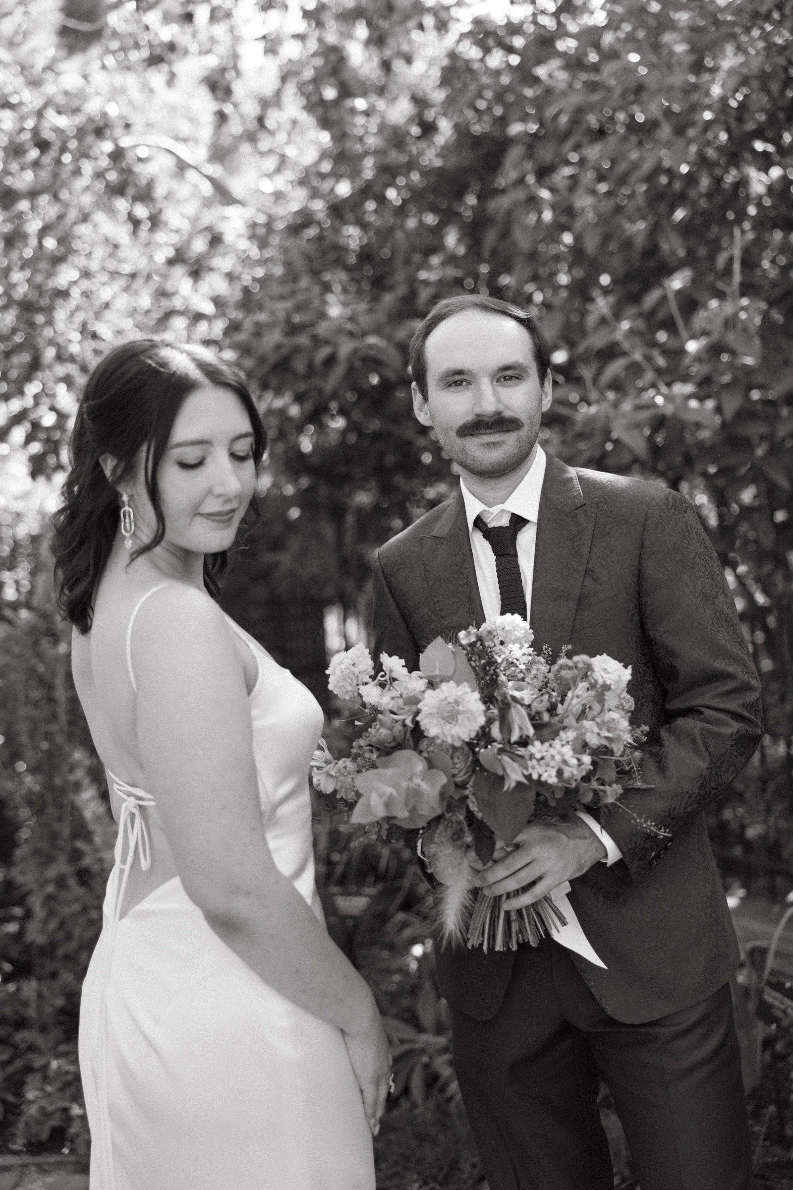 Black and white photo of a man in a suit holding a bouquet of flowers, standing next to a woman in a wedding dress, outdoors with trees in the background.