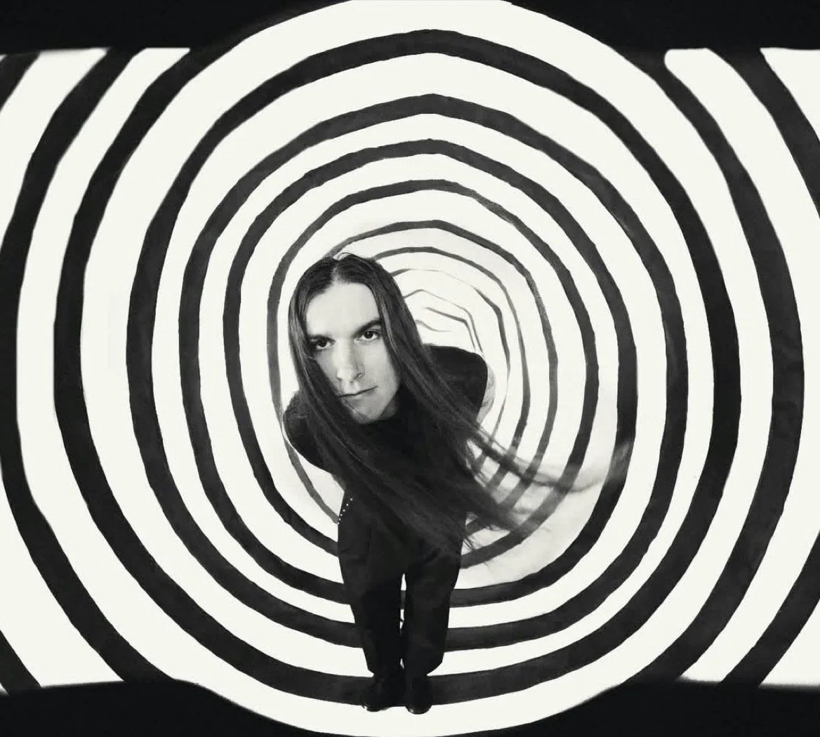 A woman with long dark hair standing in a tunnel with black and white concentric oval patterns creating a hypnotic optical illusion.