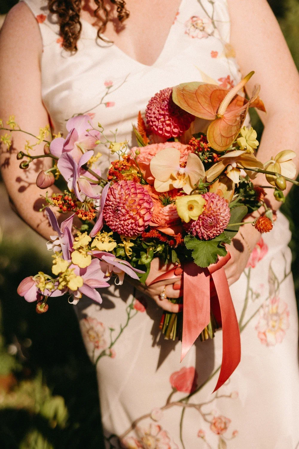 A person in a cream-colored floral dress holding a large bouquet of mixed colorful flowers, including pink, purple, yellow, and cream blooms, with a pink ribbon tied at the stems.