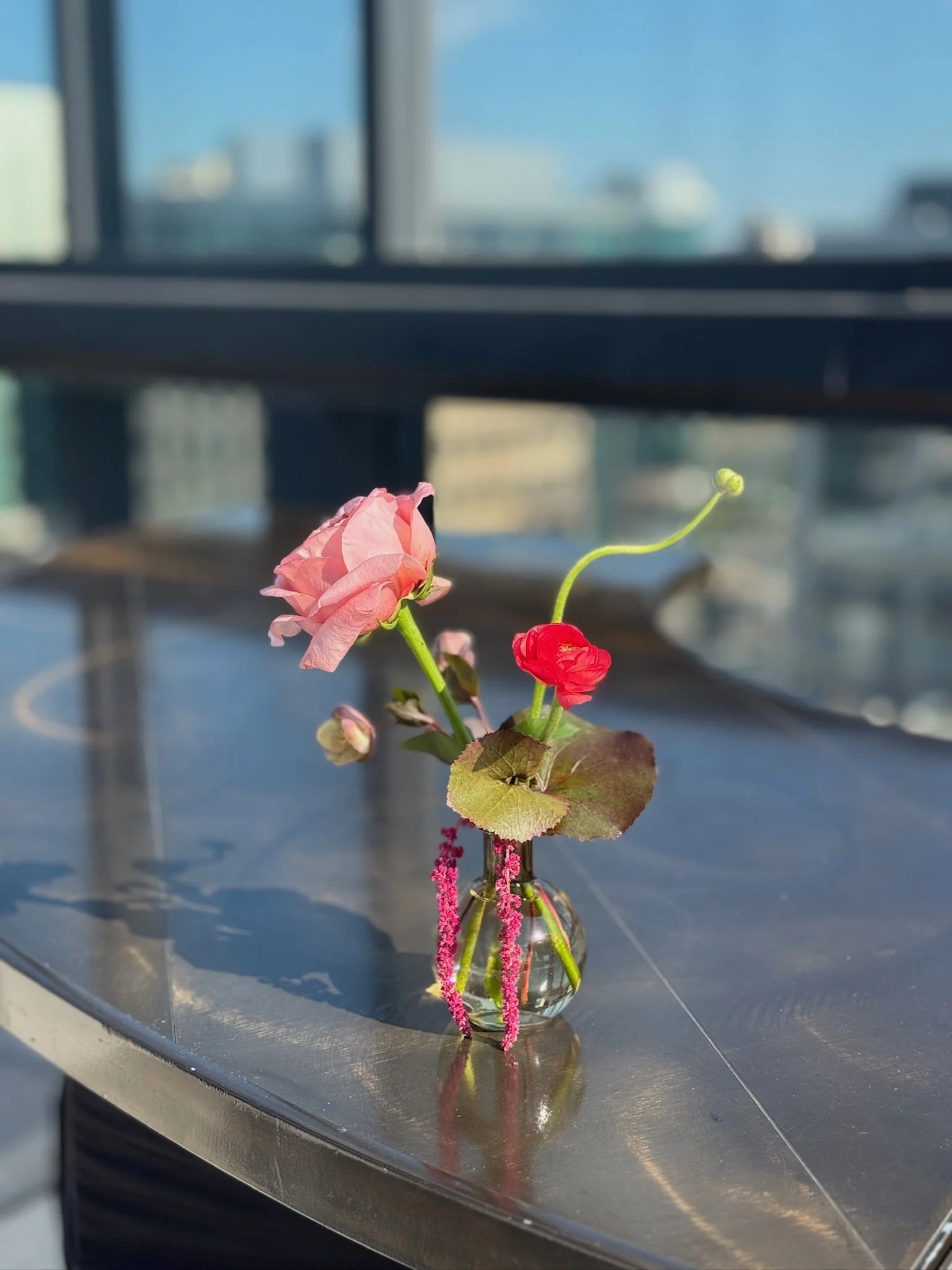 Small glass vase with pink and red flowers on a metallic table in a high-rise building, city skyline in the background.