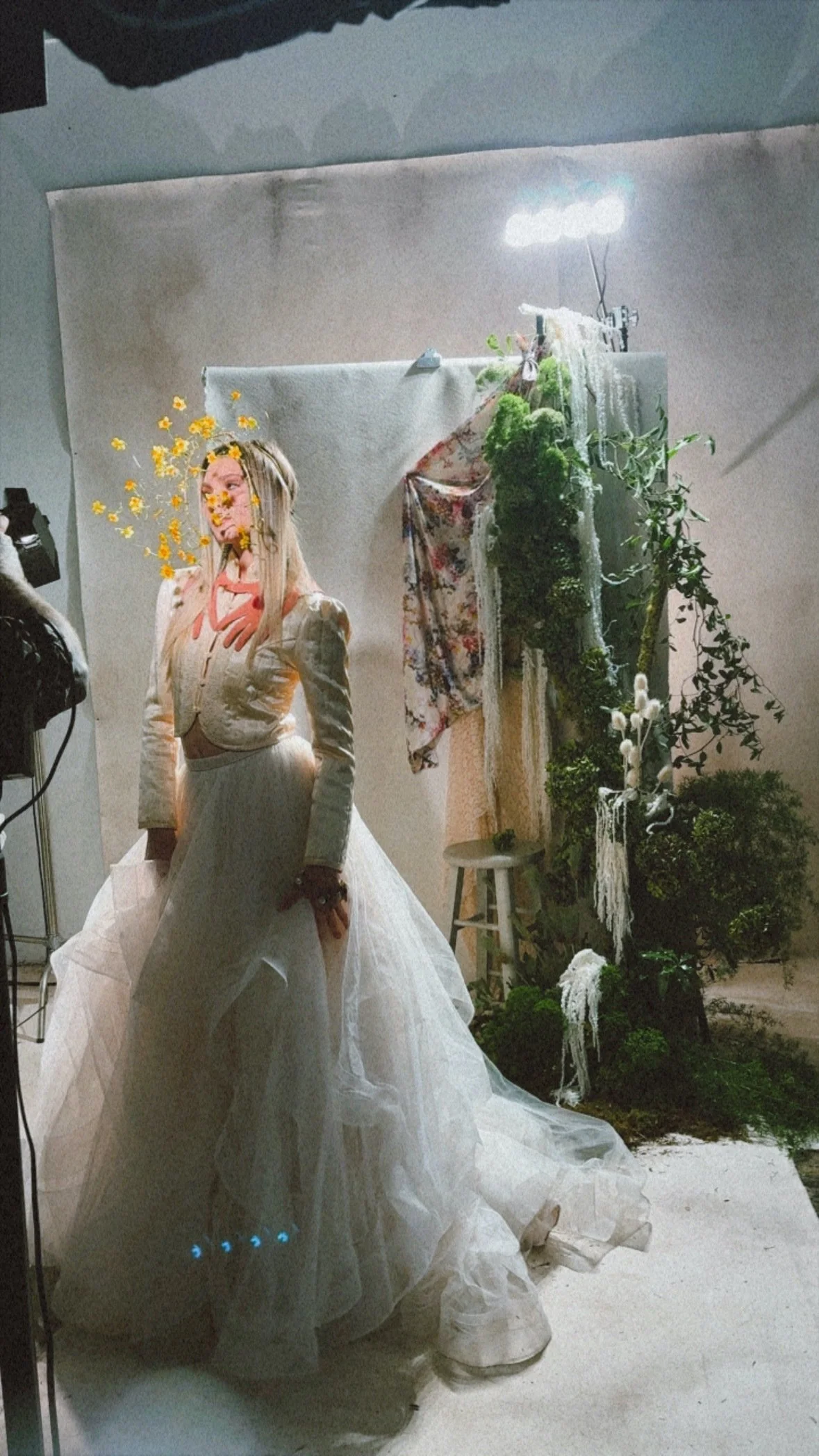A model standing in front of a photo backdrop decorated with flowers, plants, and fabric. The model is wearing a long, flowing white dress and a jacket with puffed sleeves. She is posing with her hand on her hip, and her hair is styled straight.