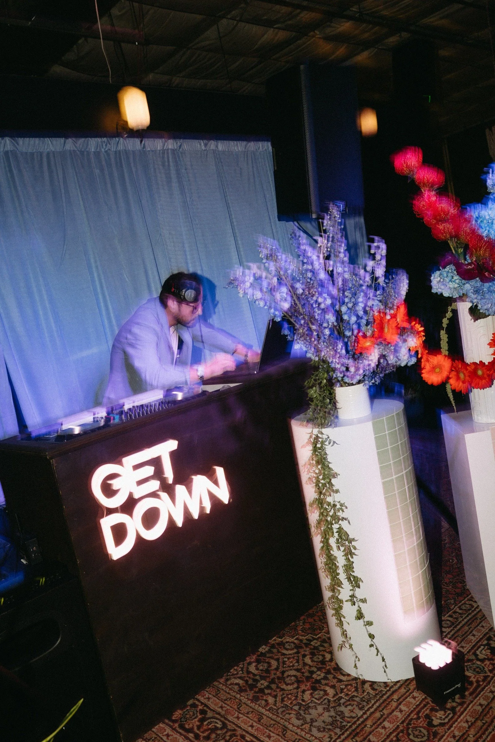 A DJ at a nightclub or party wearing headphones and working at a mixing console, with large floral arrangements and a neon sign that reads 'GET DOWN' in the foreground.