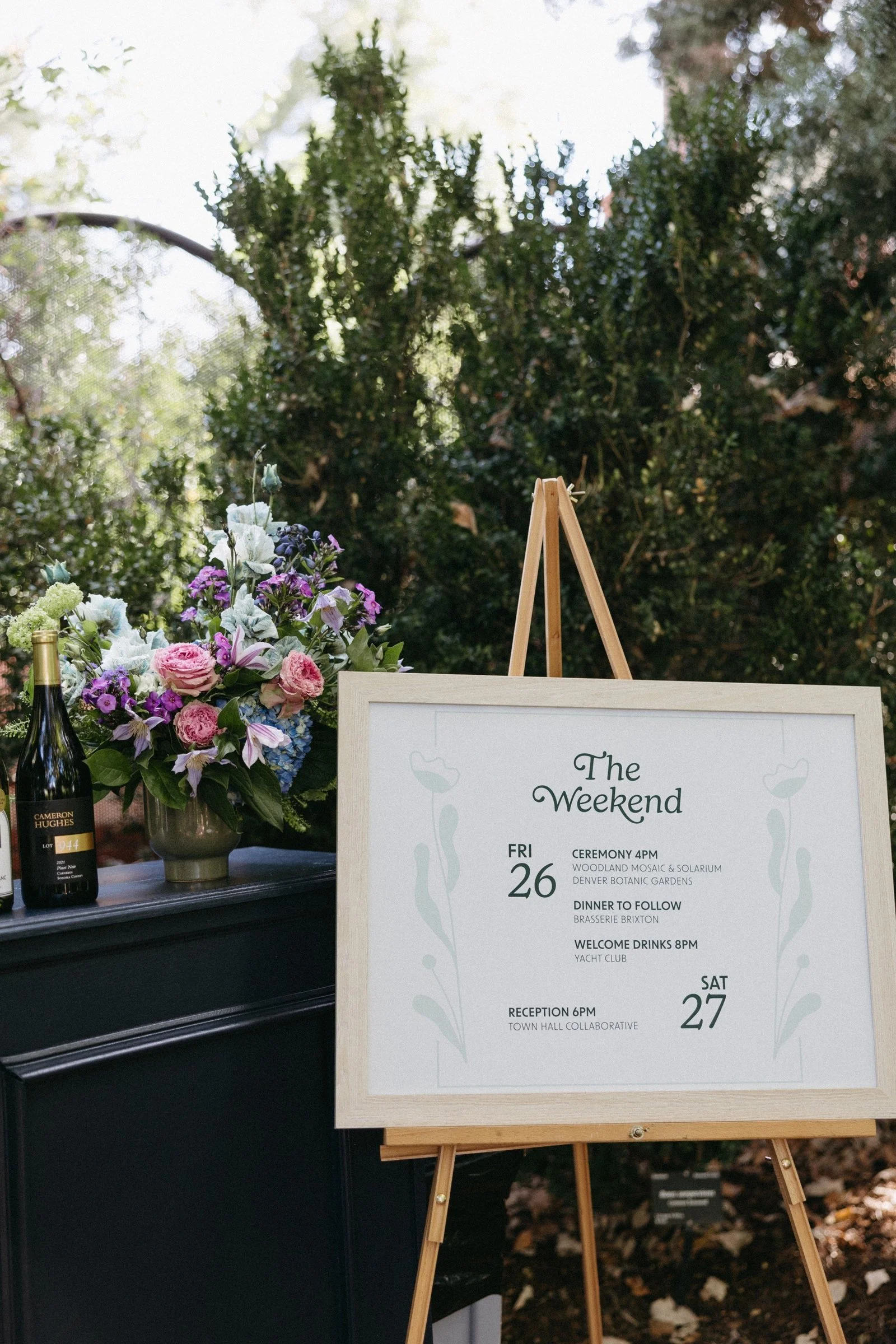 A wooden signboard detailing a weekend event schedule, positioned on an easel outdoors. It lists activities for Friday and Saturday including ceremonies, dinner, drinks, and a reception, with decorative floral graphics on the sides.