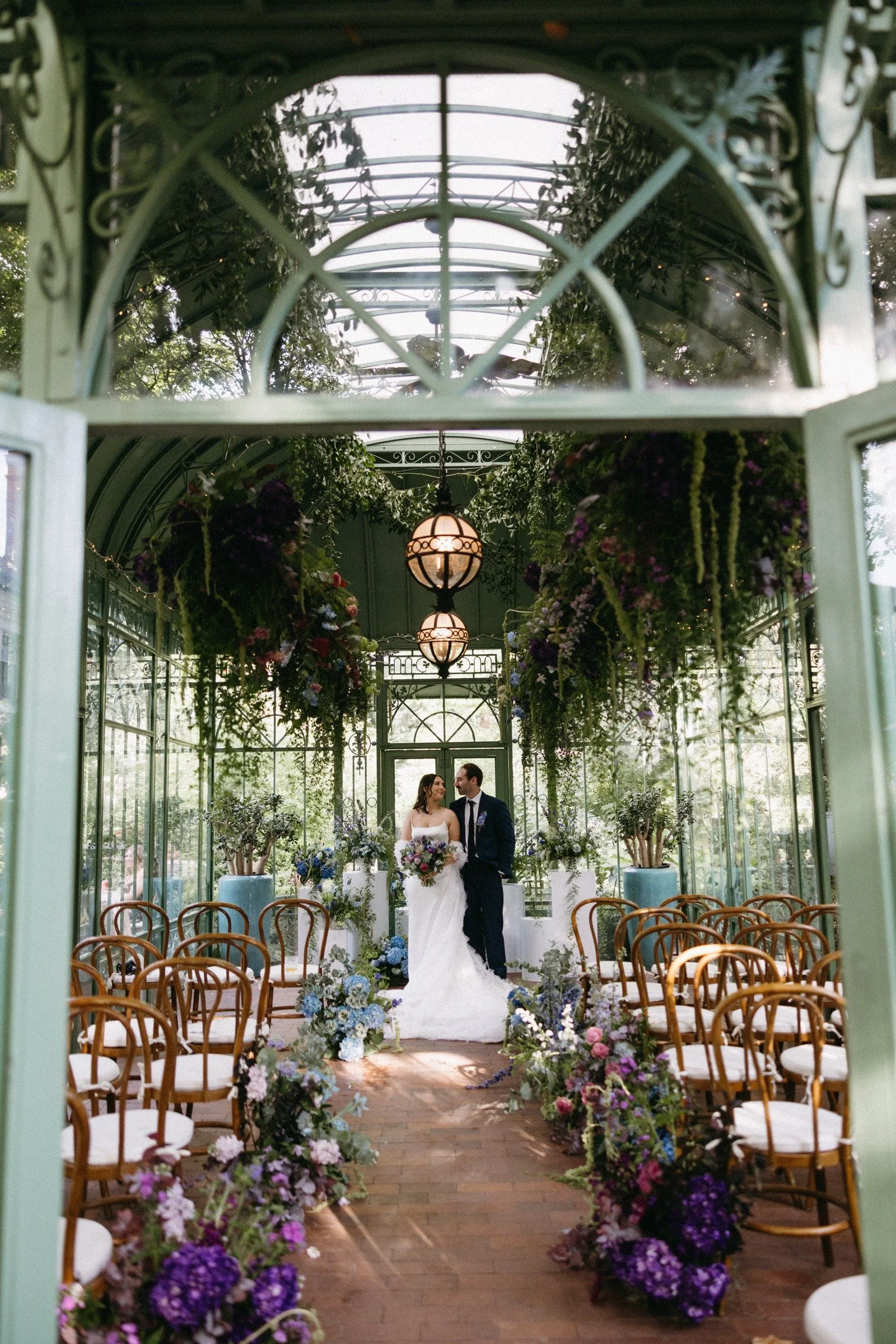 A bride and groom standing inside a glass-enclosed garden wedding venue decorated with flowers and greenery, with rows of chairs on either side for guests.