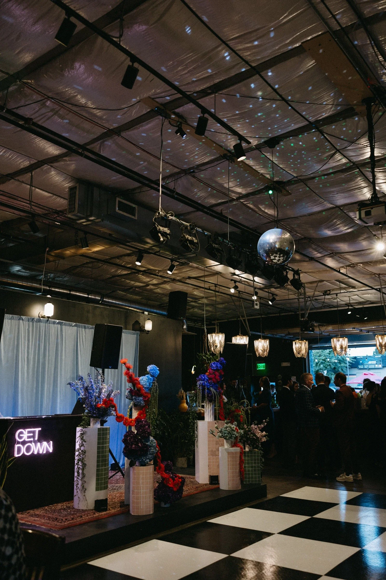 Interior of a decorated event venue with floral arrangements, a neon sign that says 'Get Down,' and a group of people socializing. The ceiling features a disco ball and colorful light effects.
