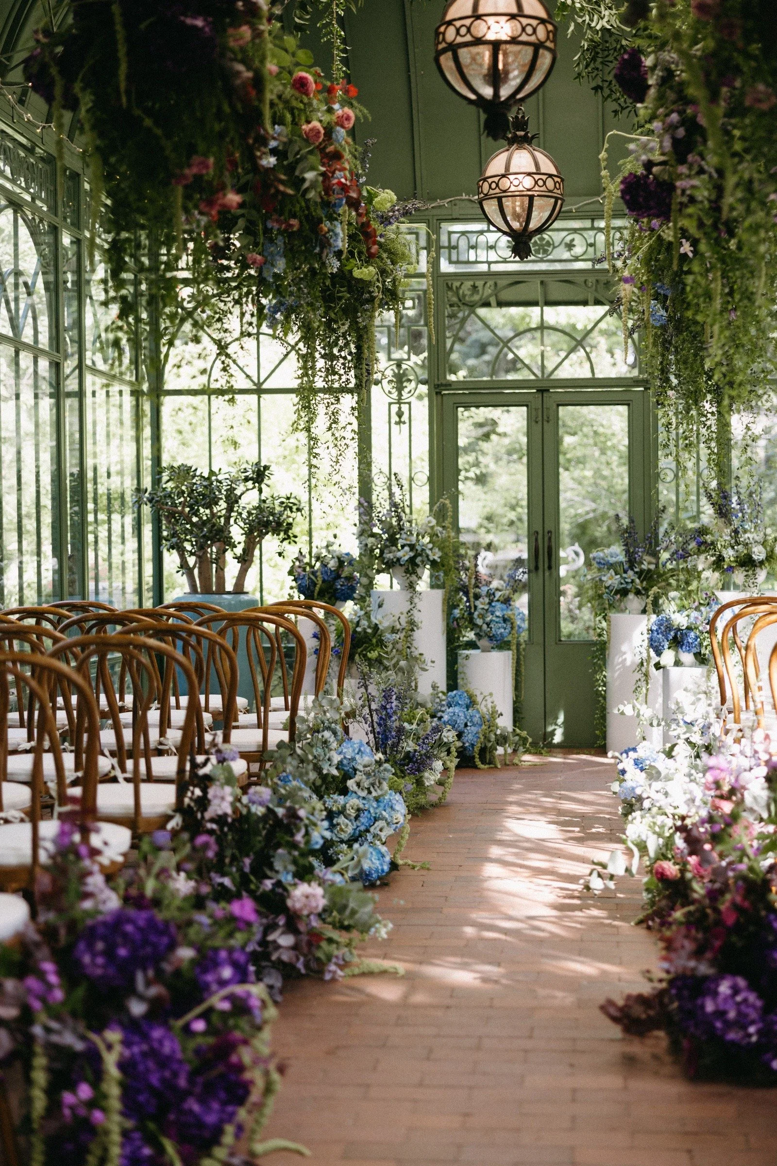A greenhouse wedding aisle decorated with lush floral arrangements in shades of purple, blue, white, and pink, with wooden chairs lining the sides and hanging greenery from the ceiling. Large lantern-style lights hang from above, and glass doors and 