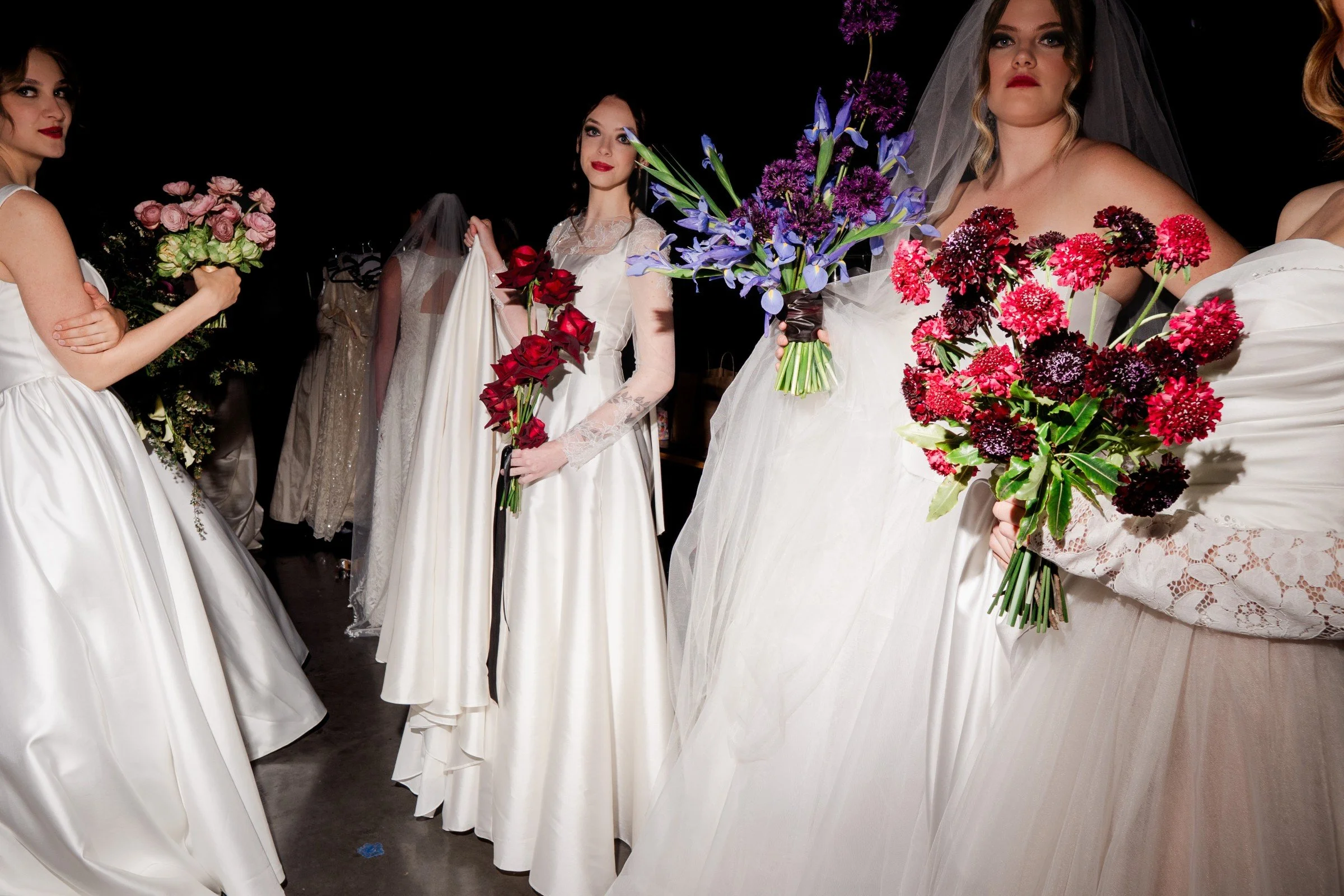 Group of women wearing wedding dresses holding colorful bouquets, standing together against a dark background.