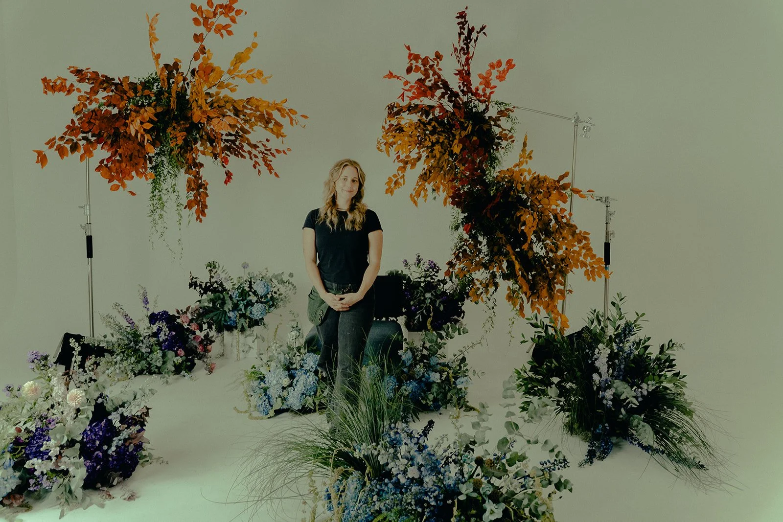 A woman standing in a studio surrounded by various colorful floral arrangements and large autumn-colored foliage displays against a neutral background.