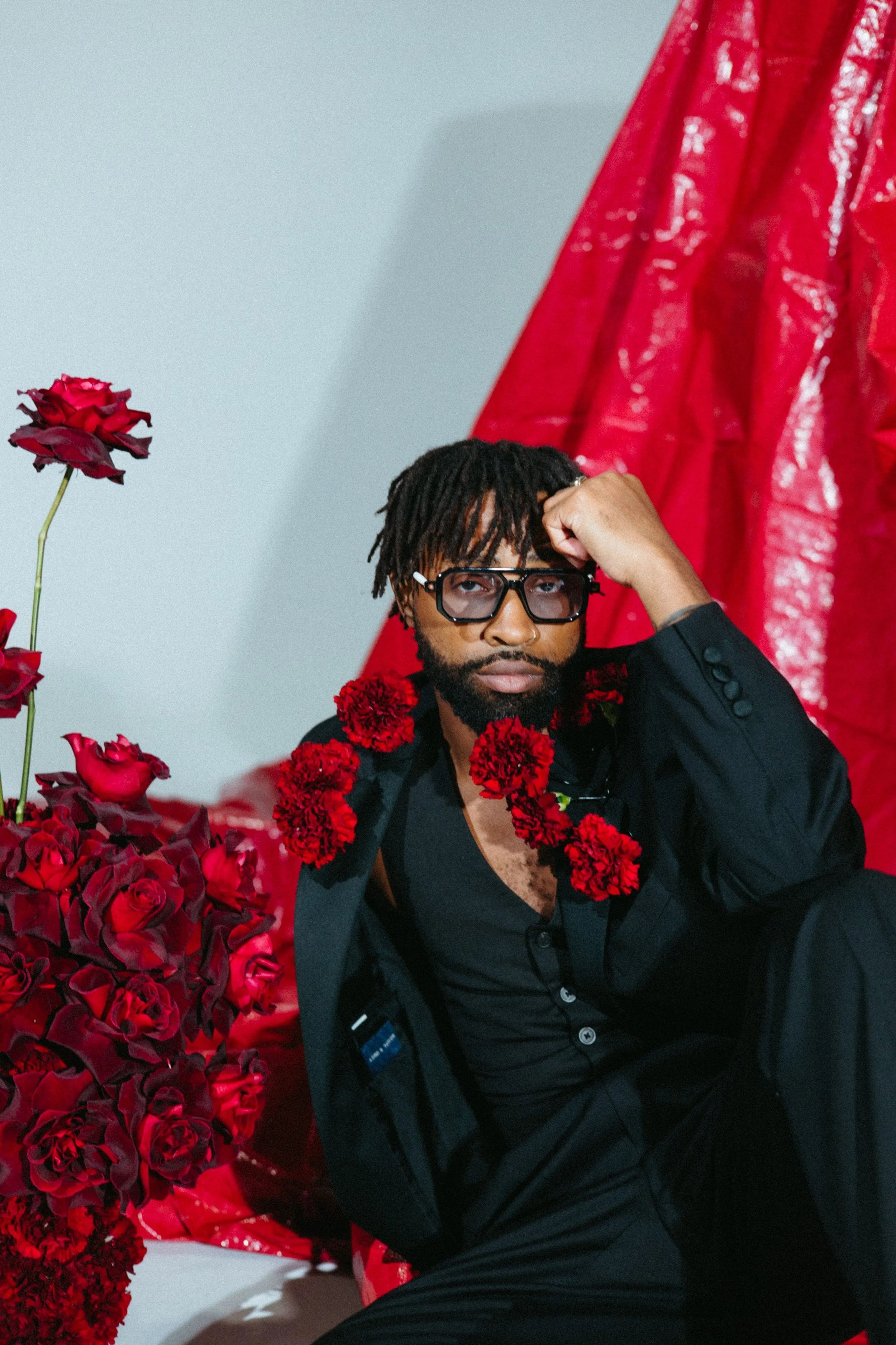A man with dreadlocks and glasses sitting with red flowers around his neck, surrounded by red roses and a red textured backdrop.