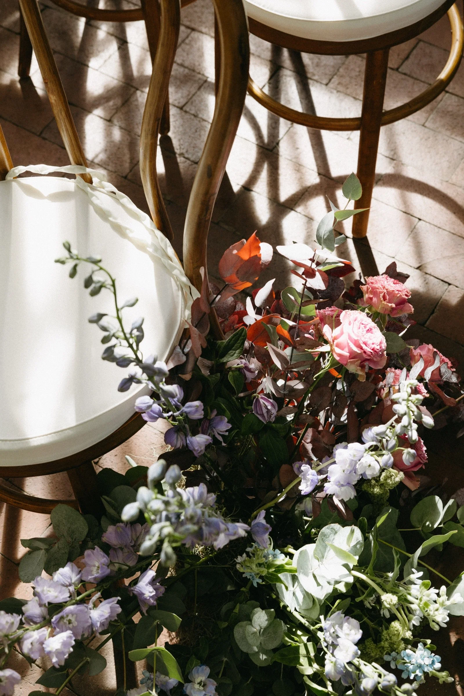 A bouquet of pink, purple, and white flowers on the floor next to wooden chairs in a sunlit room.