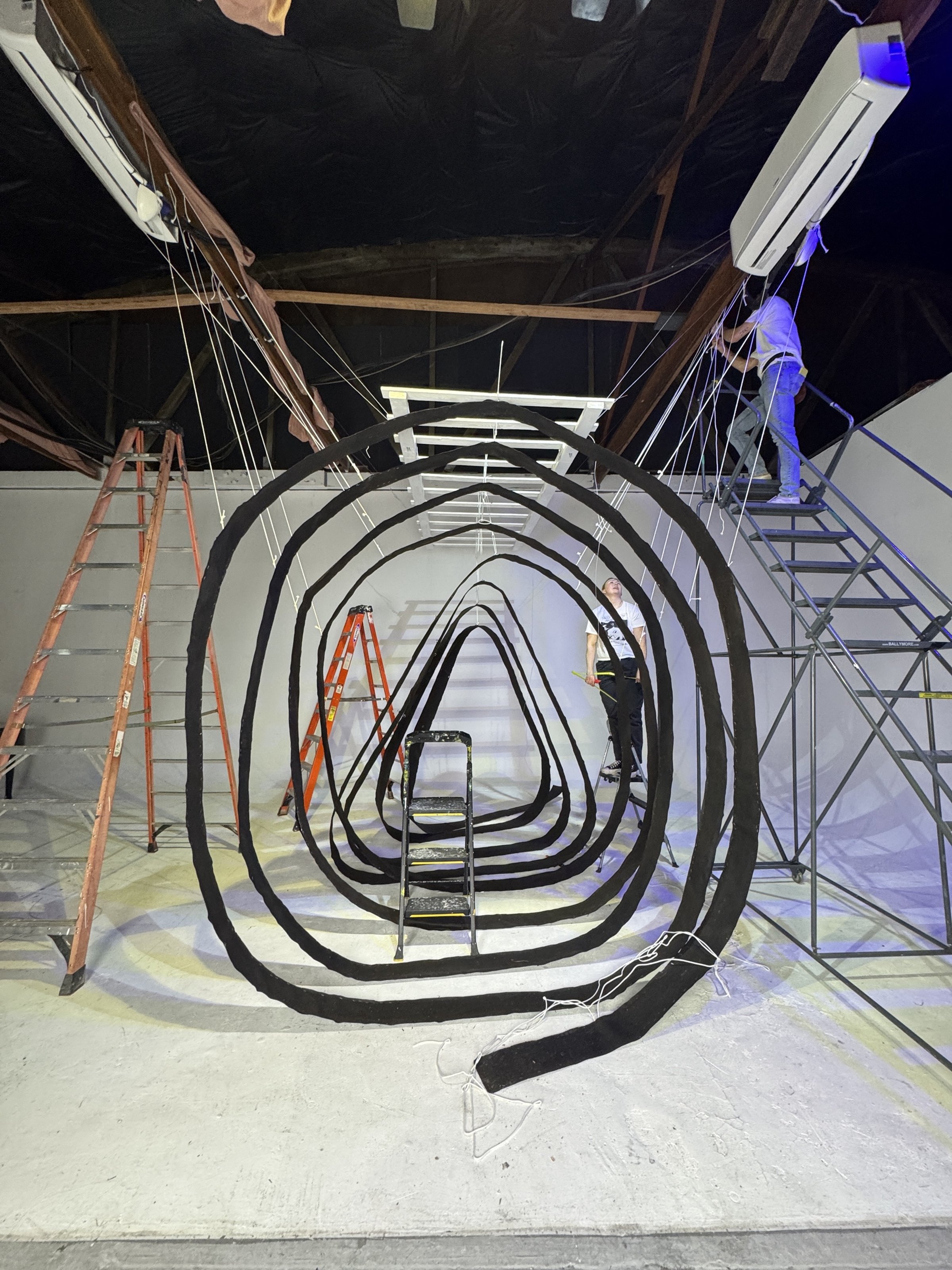 People setting up an art installation with concentric black oval shapes, ladders, and scaffolding inside a room with a black ceiling and white walls.