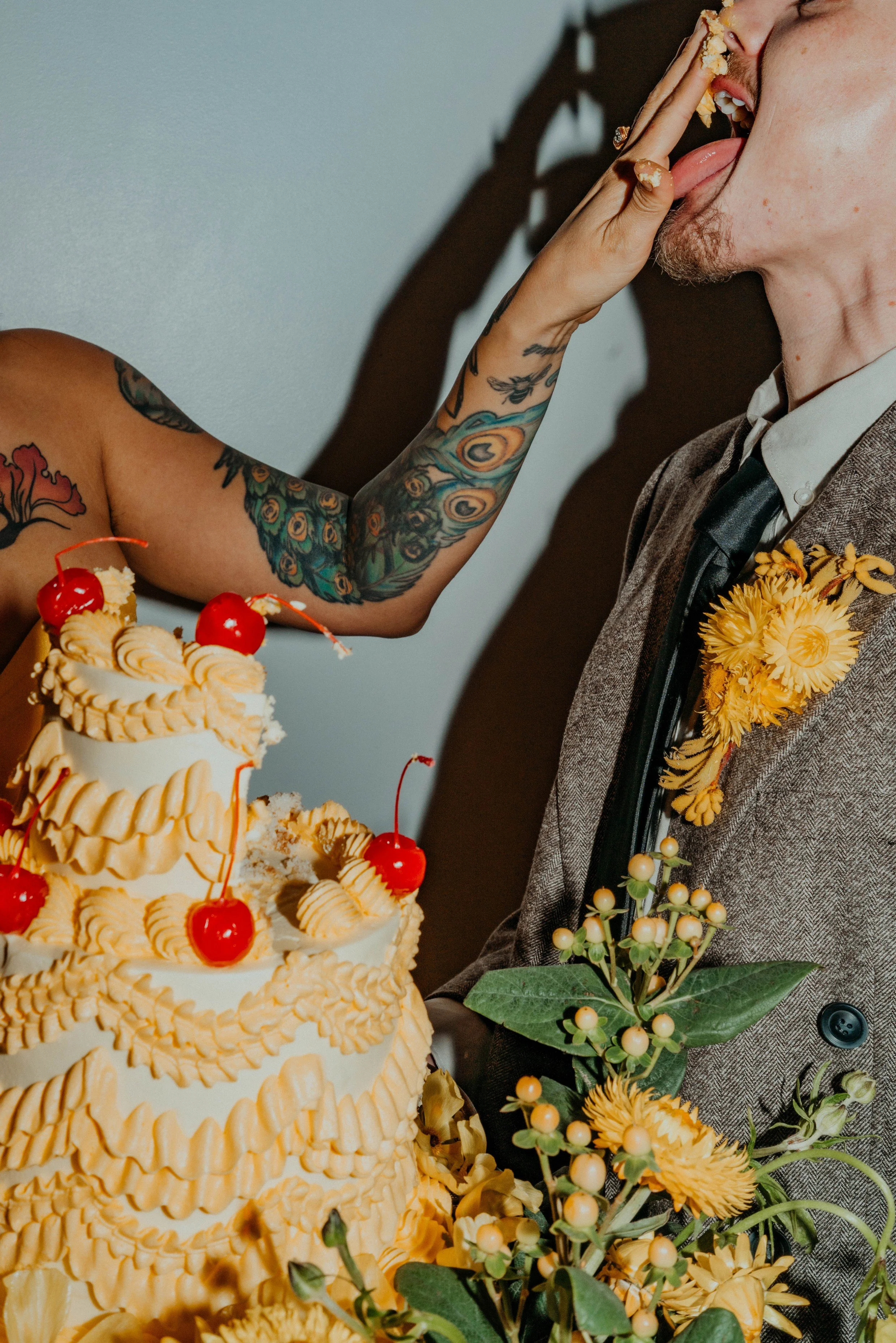 A groom with a patch of facial hair wears a brown suit with a floral boutonniere, as a bride with colorful tattoos feeds him a piece of cake. The cake is decorated with cherries and cream, and there are floral decorations nearby.