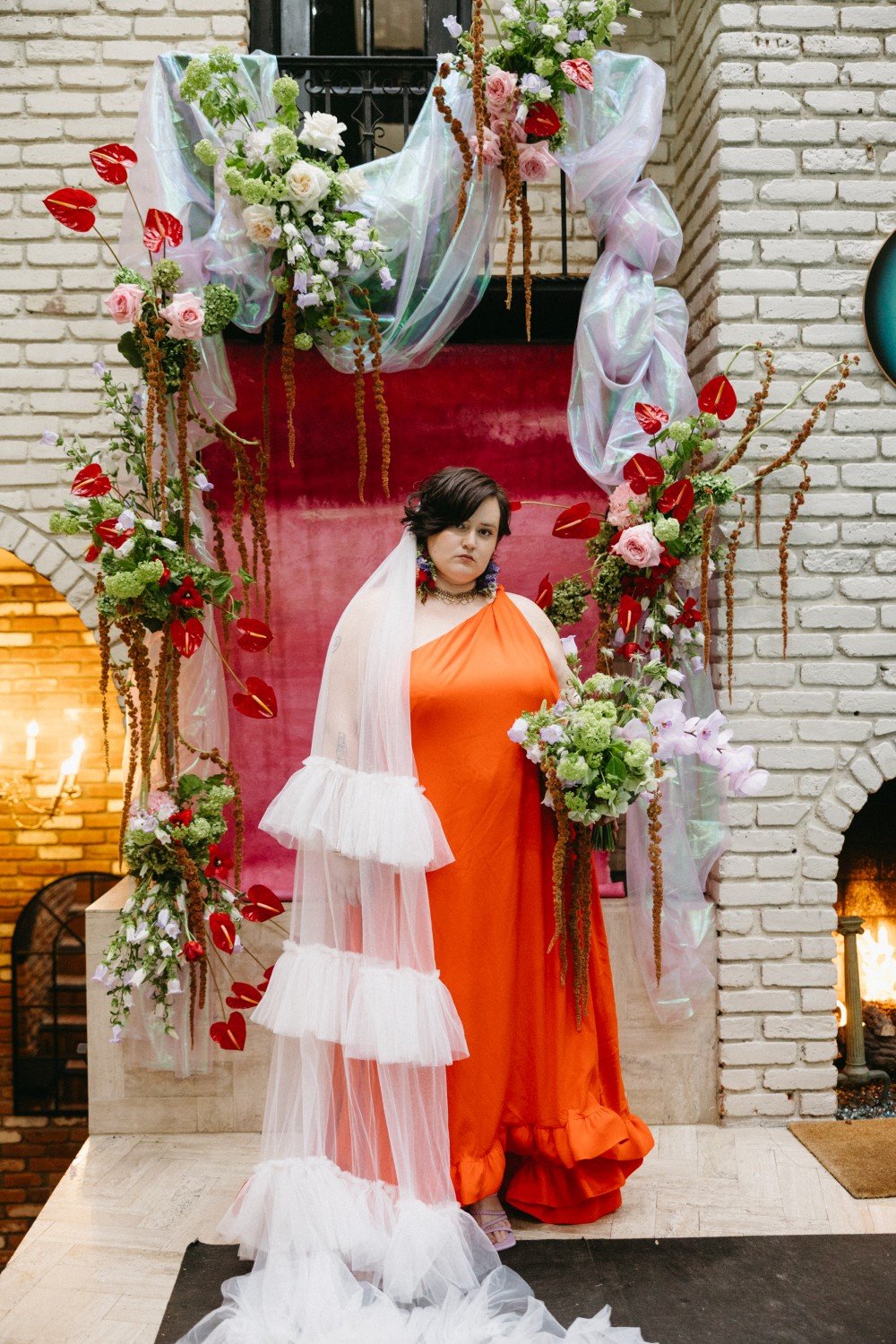 A person with short dark hair wearing an orange dress stands amidst a floral arrangement with pink, white, and red flowers, greenery, and decorative elements in front of a white brick wall. There is a lit fireplace in the background.