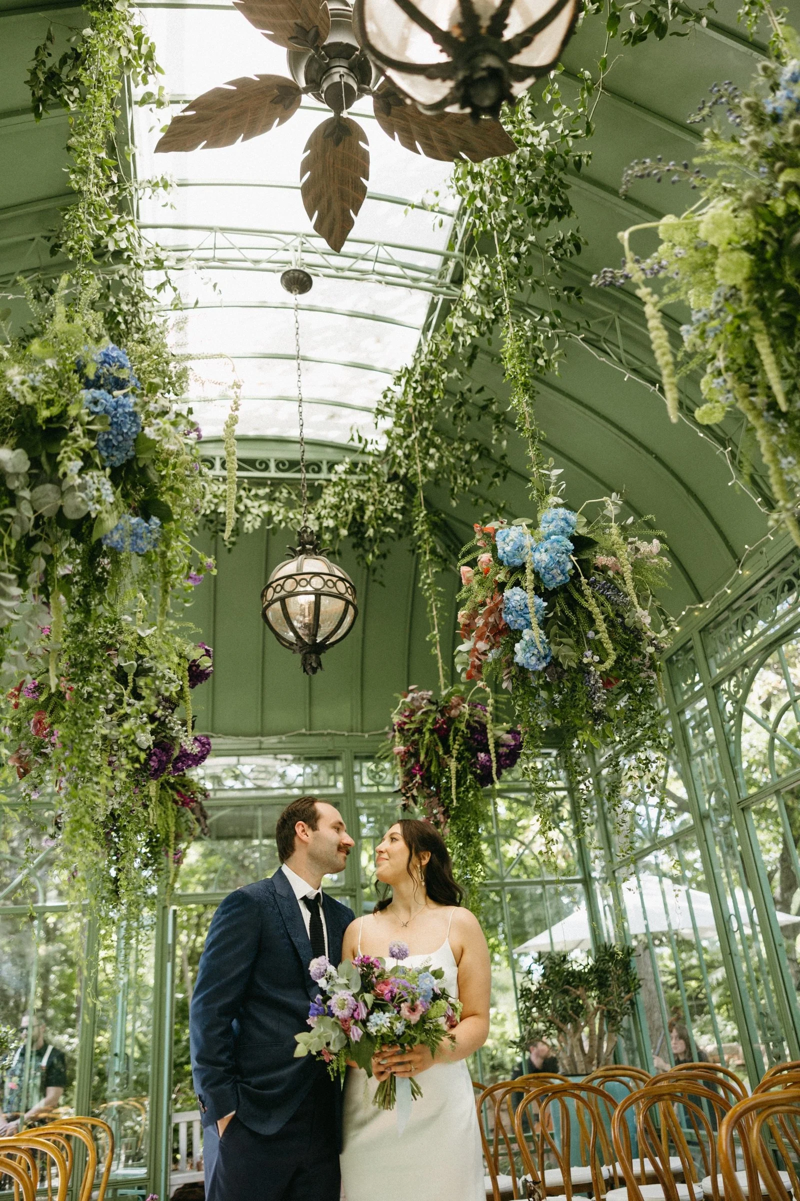 A bride and groom standing close together inside a green, glass-enclosed conservatory decorated with hanging flowers and lush greenery, with chairs arranged around them for a wedding ceremony.