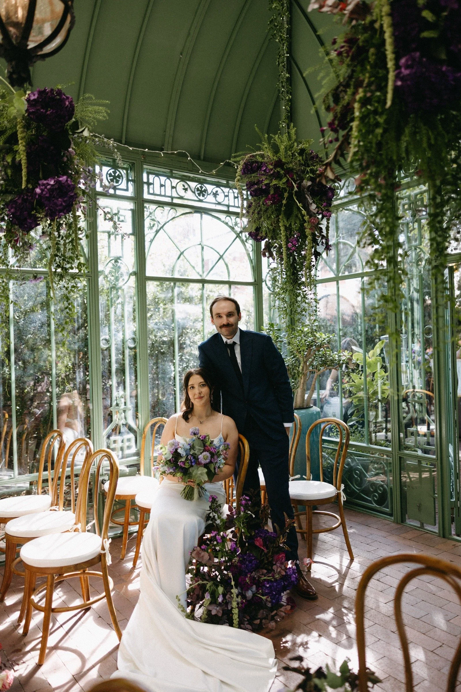 A bride and groom posing inside a glass greenhouse with floral decorations, sunlight streaming through large windows, and wooden chairs arranged around.