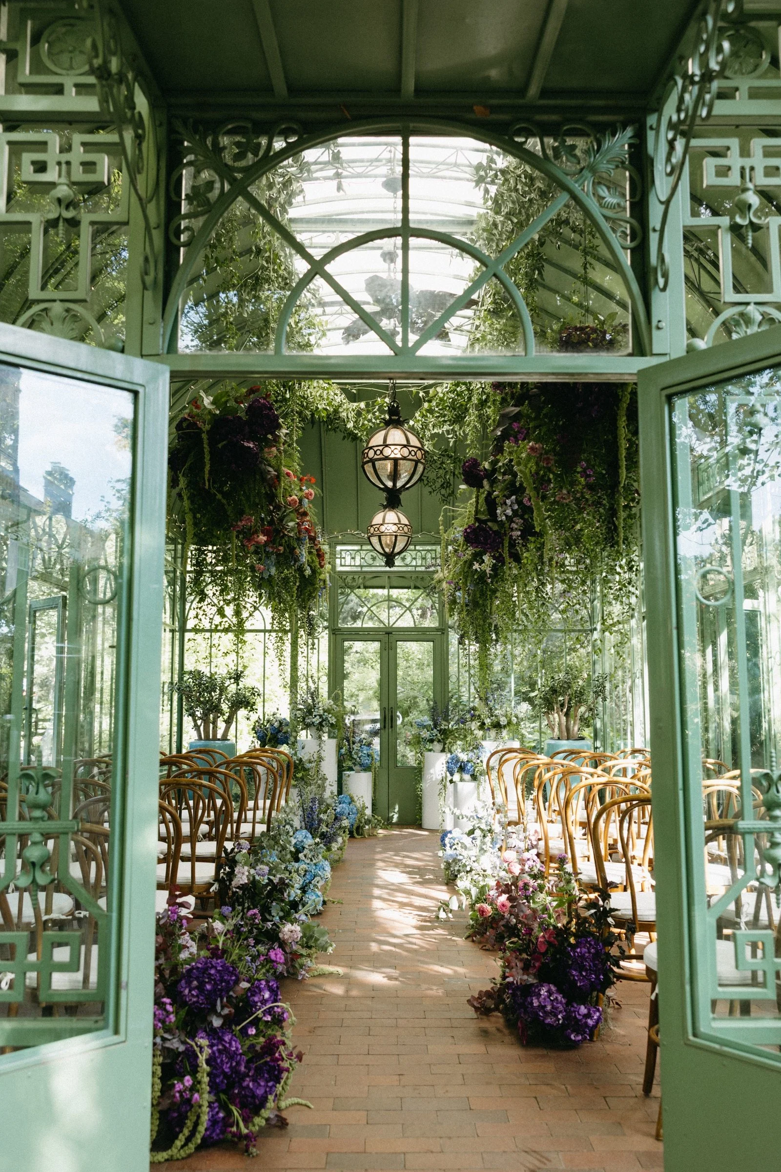 A sunlit glass greenhouse with green metal framing, decorated with hanging and potted flowers along the aisle, leading to double glass doors.