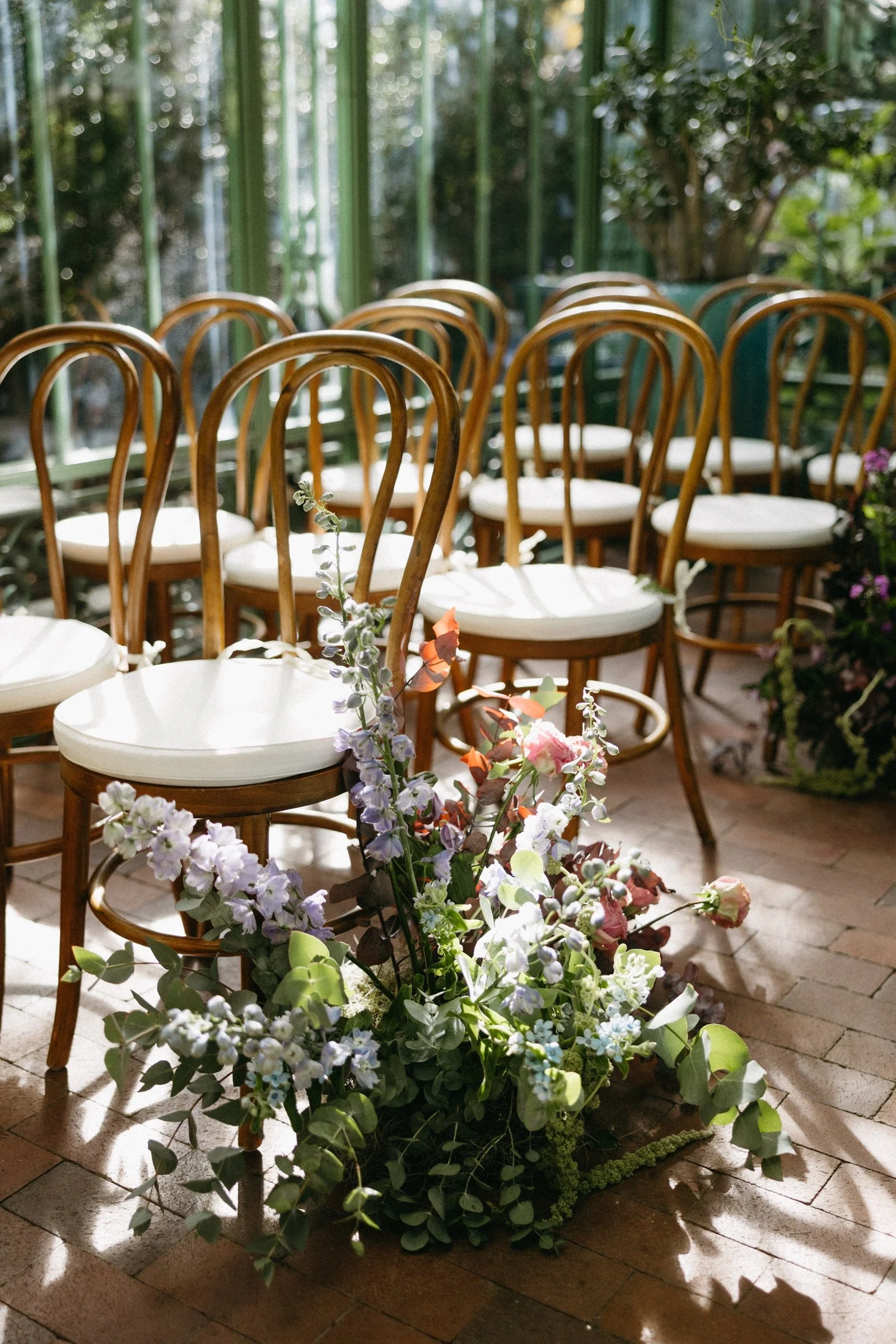 Rows of wooden chairs with white cushions arranged in an indoor space decorated with flowers and greenery.