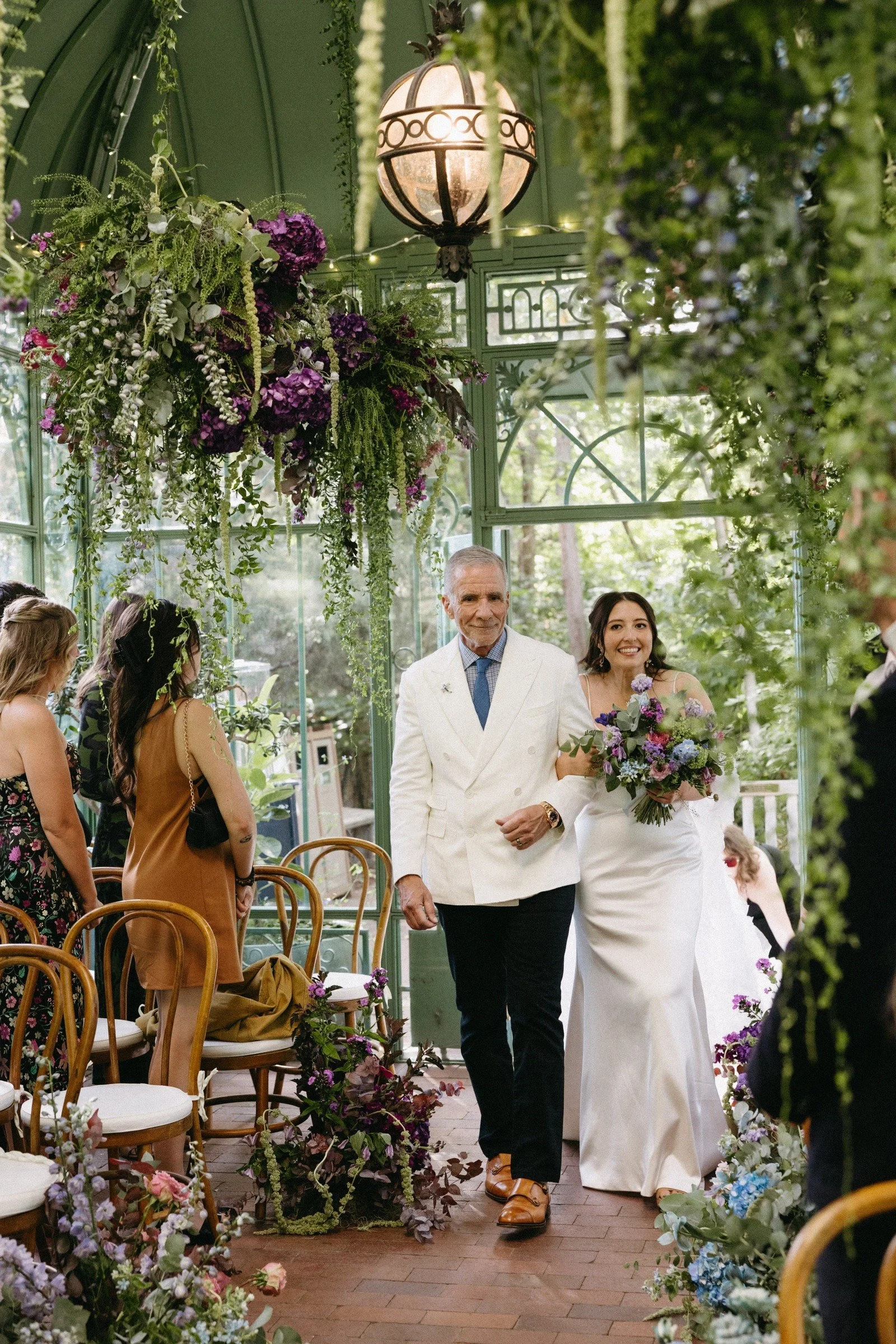 A bride in a white gown walking down the aisle with an older man, likely her father, in a light-colored suit, at a wedding ceremony decorated with lush greenery and purple flowers in a glass greenhouse setting.