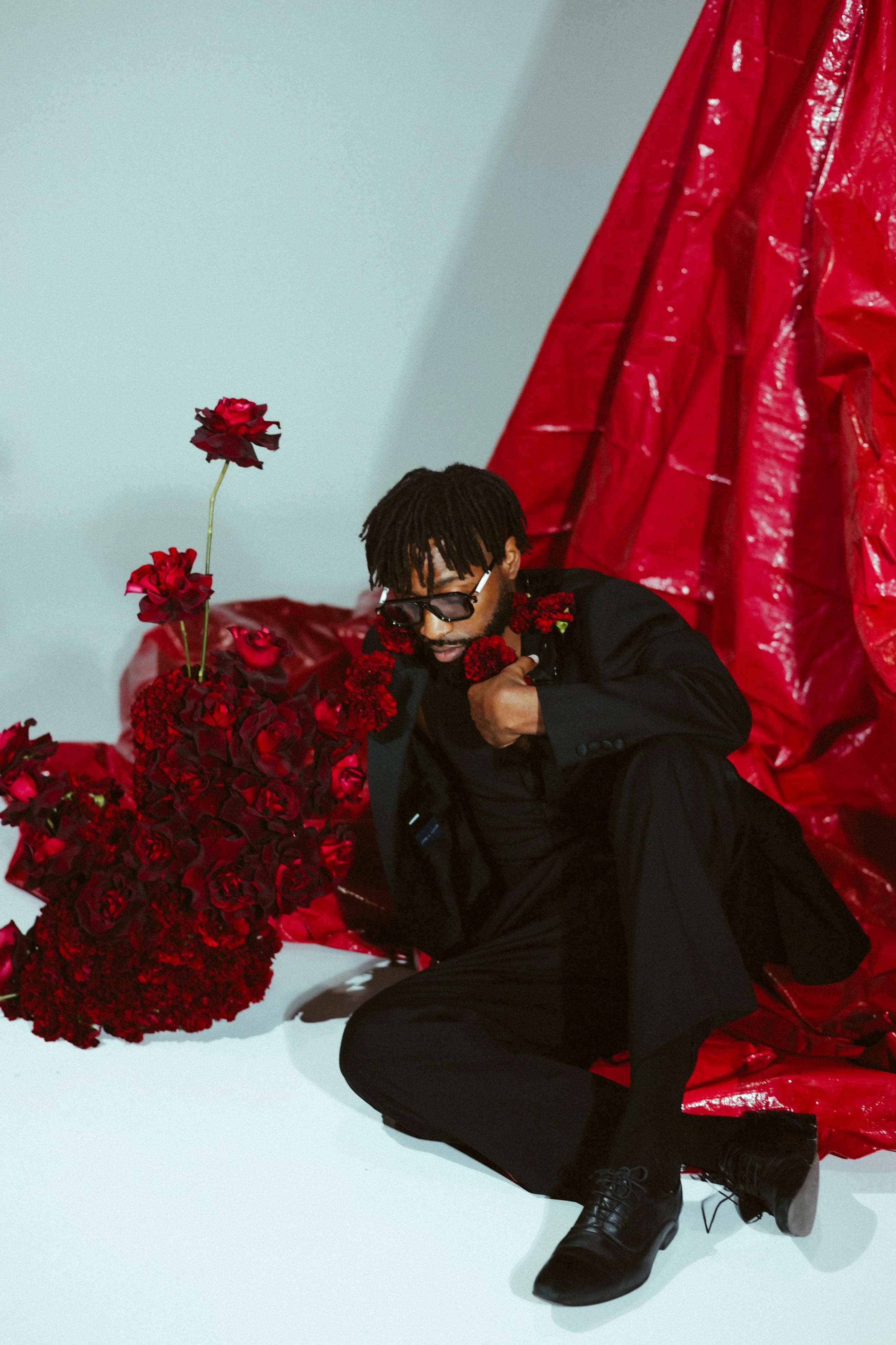 a Black male model in a dark suit against a background of white with a red backdrop and red carnations and roses