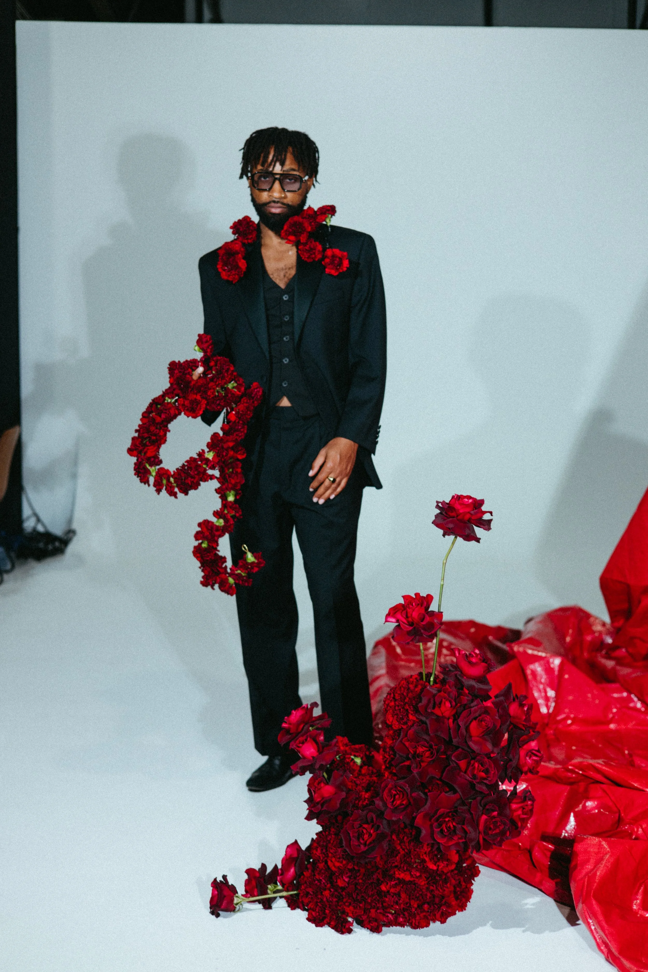 a Black male model in a dark suit against a background of white with a red backdrop and red carnations and roses