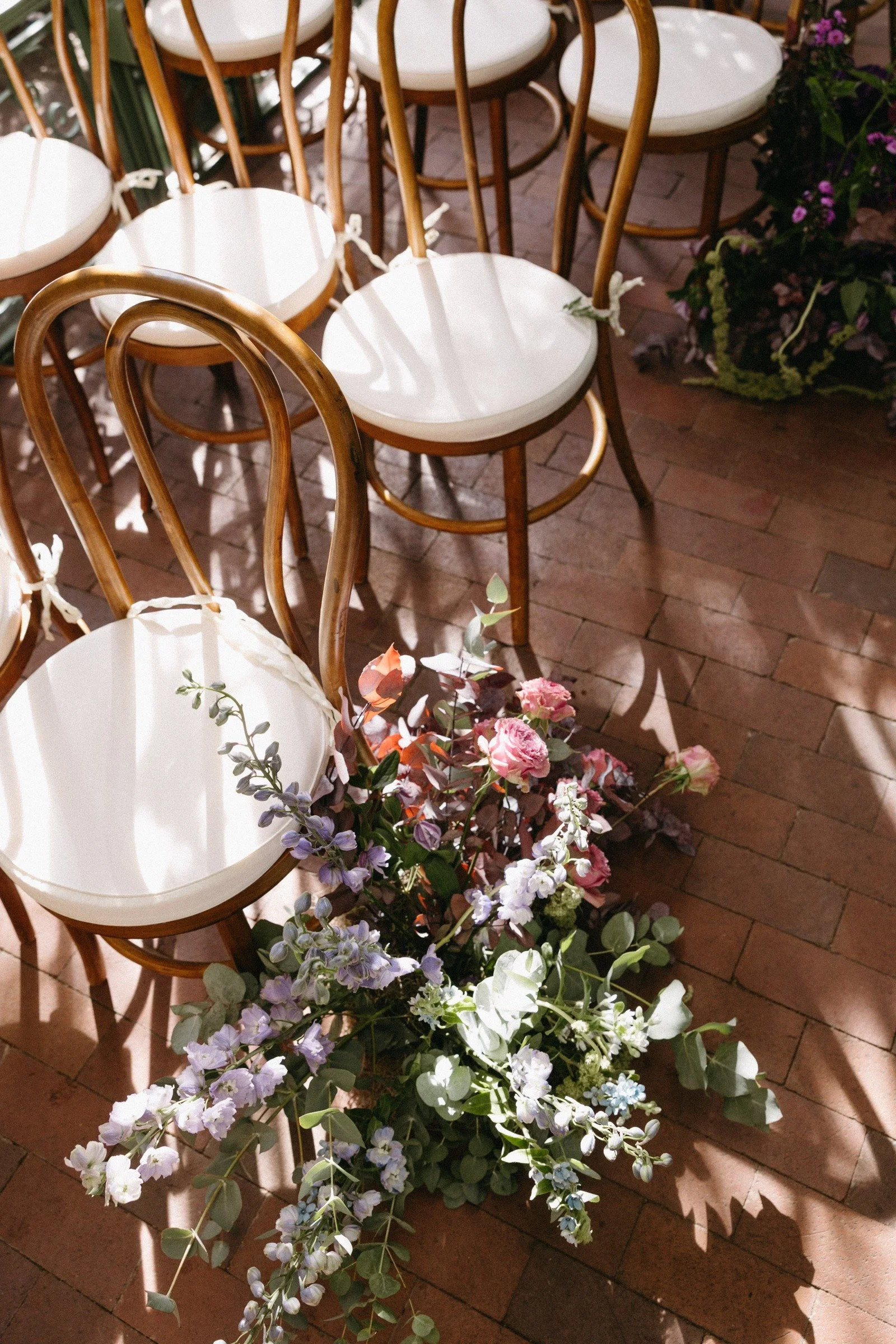 Multiple white-cushioned chairs with wooden frames arranged in a semi-circle, with a floral arrangement of pink, purple, and white flowers on a brick floor, in a setting with natural sunlight.