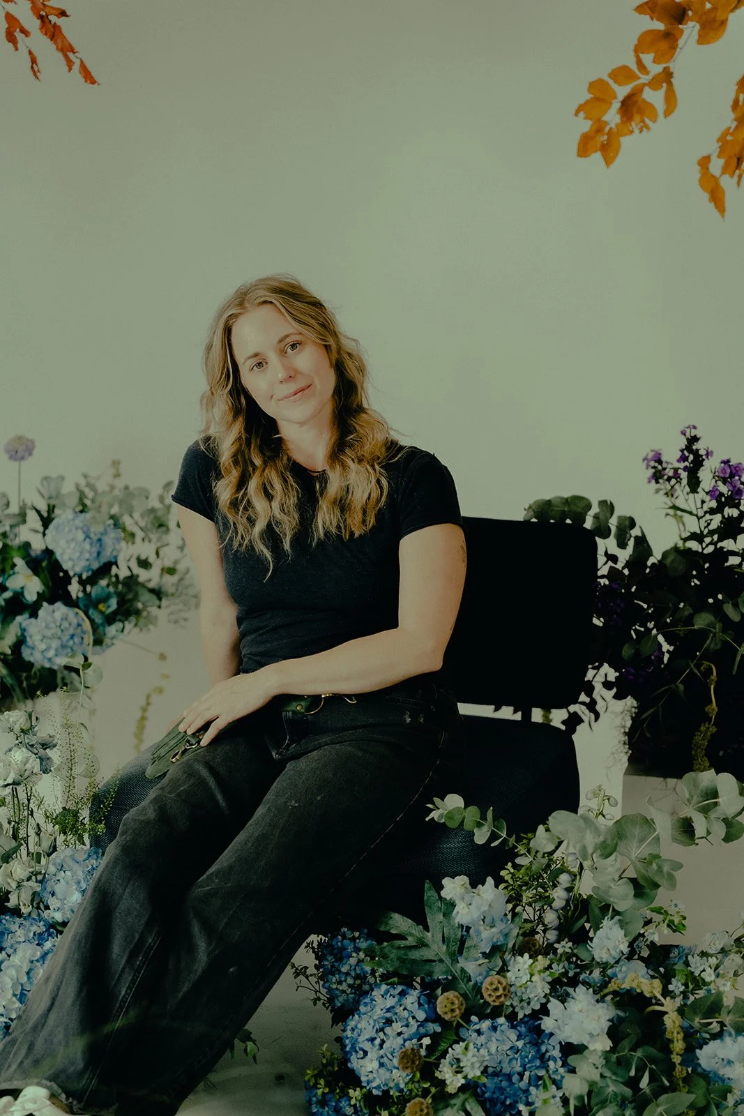 A woman with wavy, blonde hair sitting on a black chair surrounded by various colorful flowers, including blue, purple, and white, against a plain light-colored background.