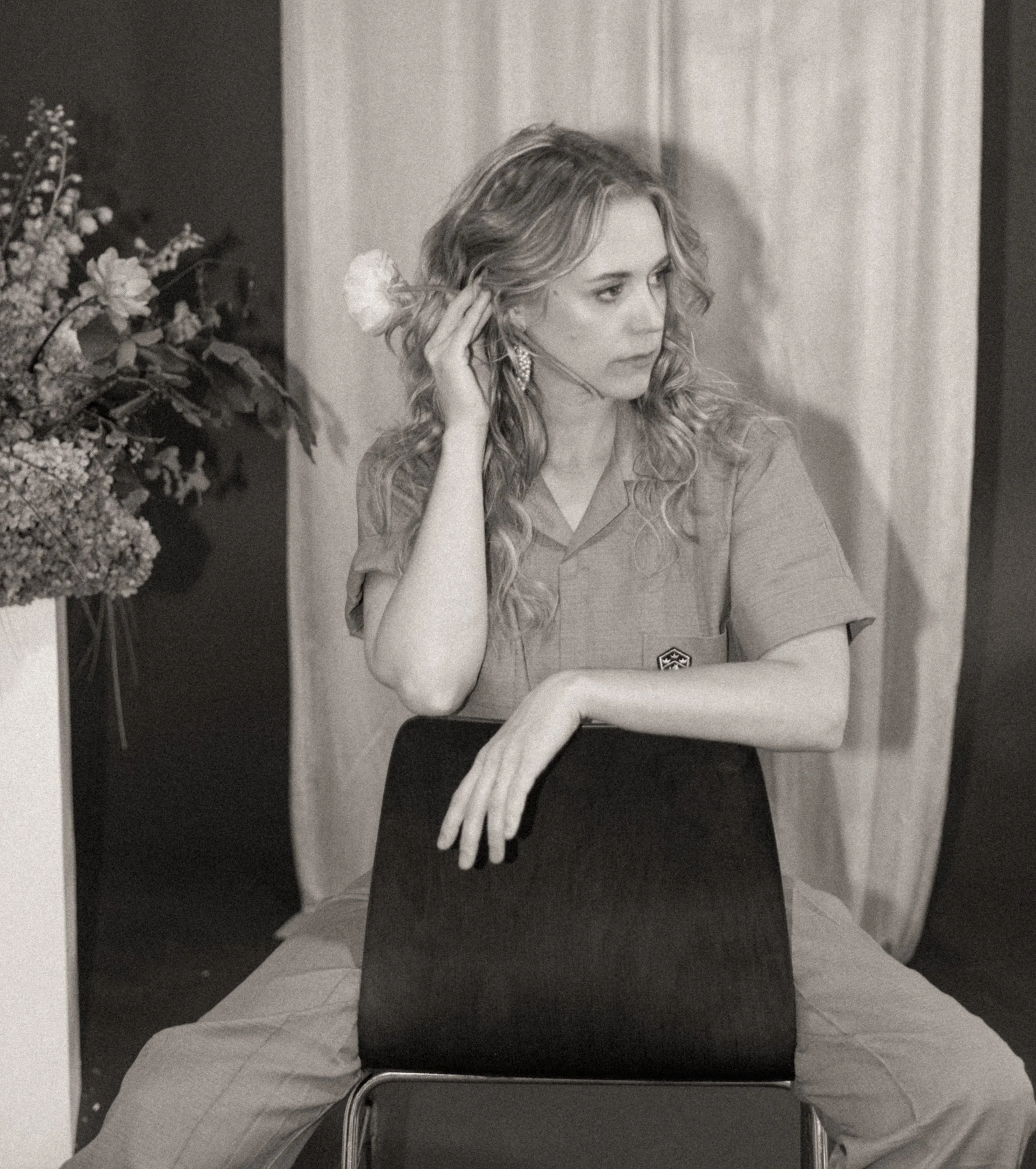 Black and white photo of a woman with curly hair, wearing earrings and a shirt with a badge, sitting backwards on a chair with one arm resting on the chair back and the other hand touching her hair, next to a bouquet of flowers.