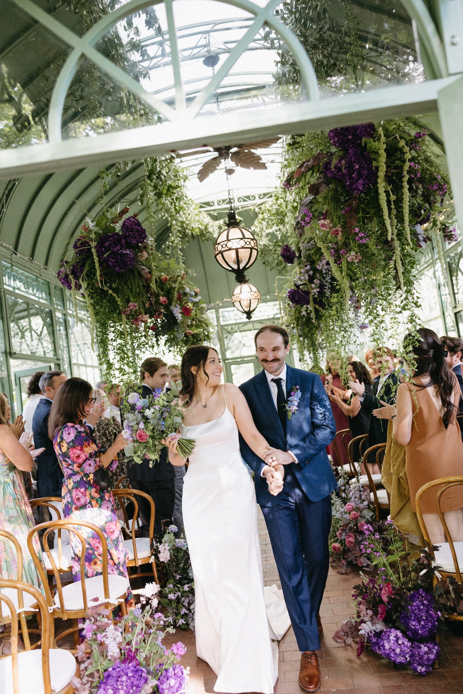 A bride and groom walking down the aisle at a wedding ceremony inside a glass greenhouse, surrounded by guests and decorated with hanging and floral arrangements.