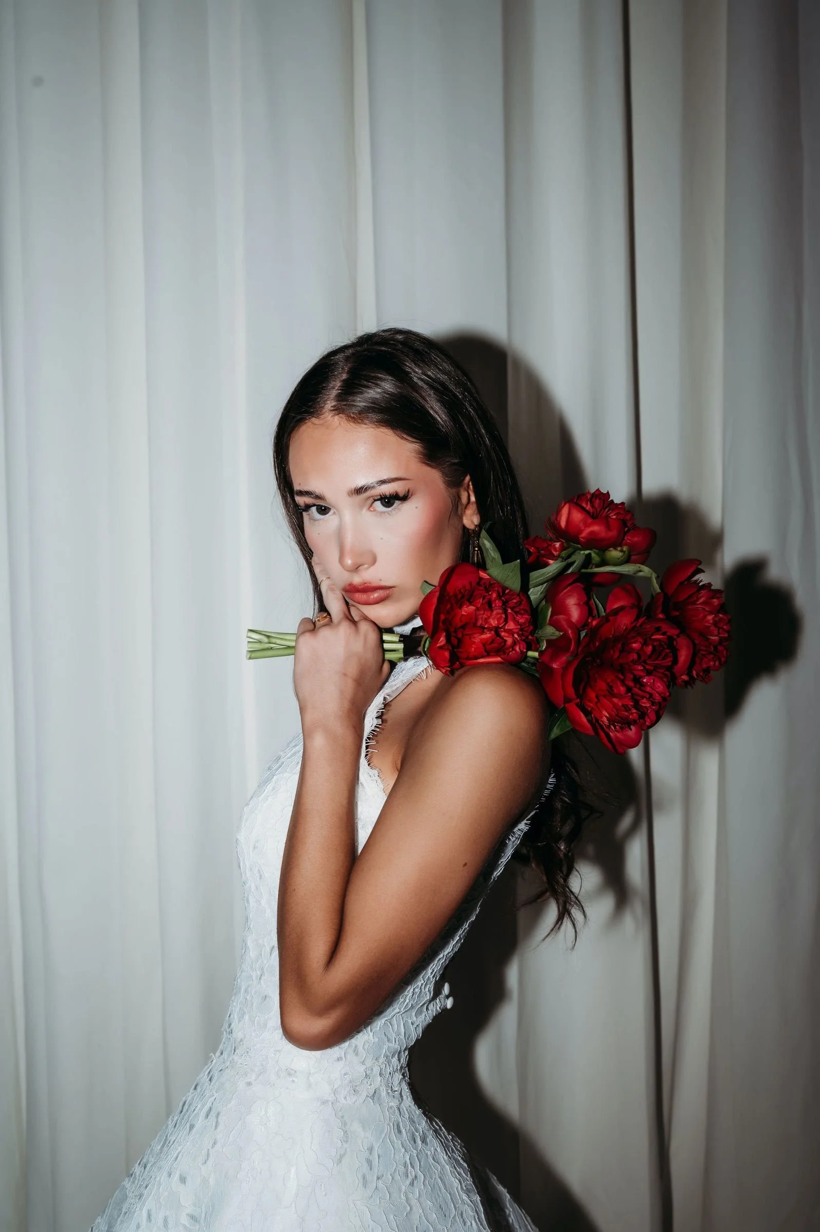 Young woman with dark brown hair and makeup dressed in a white lace wedding gown holding a bouquet of red roses on her shoulder, standing against a cream-colored curtain backdrop.