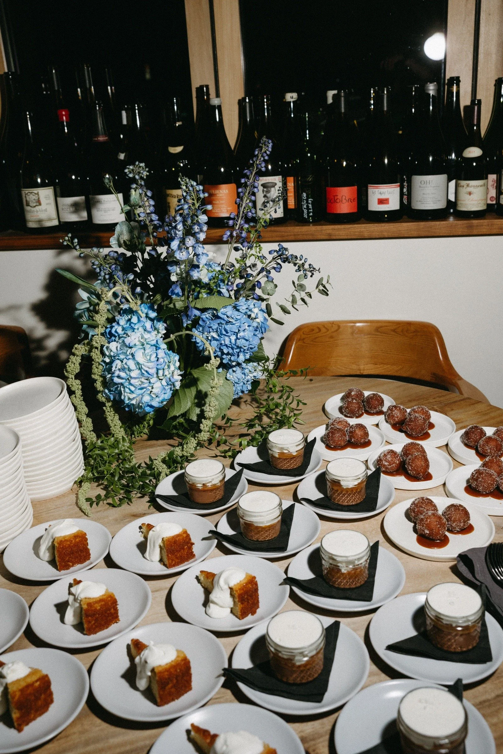 A table with plates of cake and dessert jars, decorated with a large blue and purple floral centerpiece, set against a background of wine bottles.