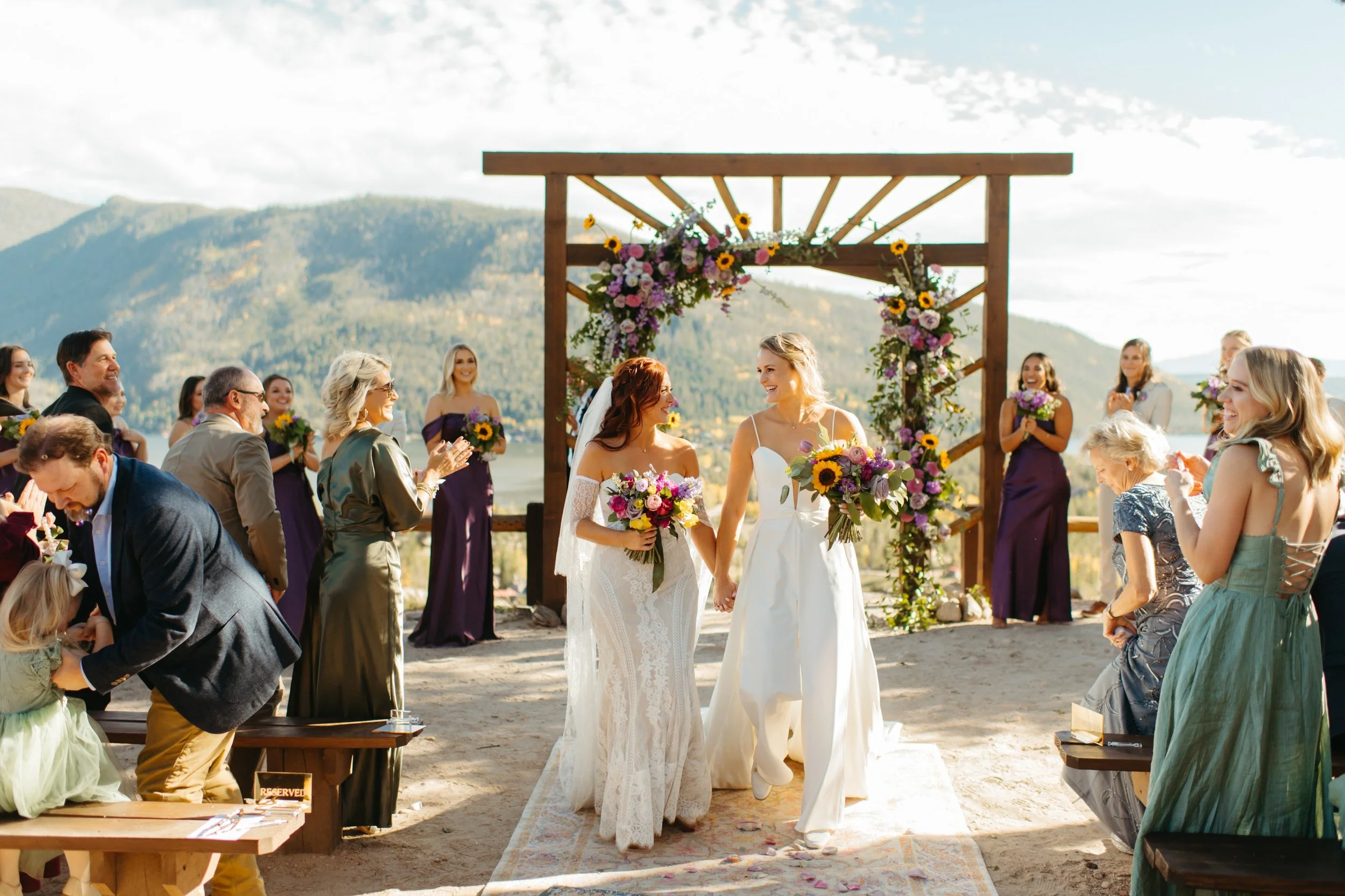 A wedding ceremony on a mountain overlook with two brides holding hands and smiling at each other, surrounded by guests and bridesmaids, with scenic mountains and sky in the background.