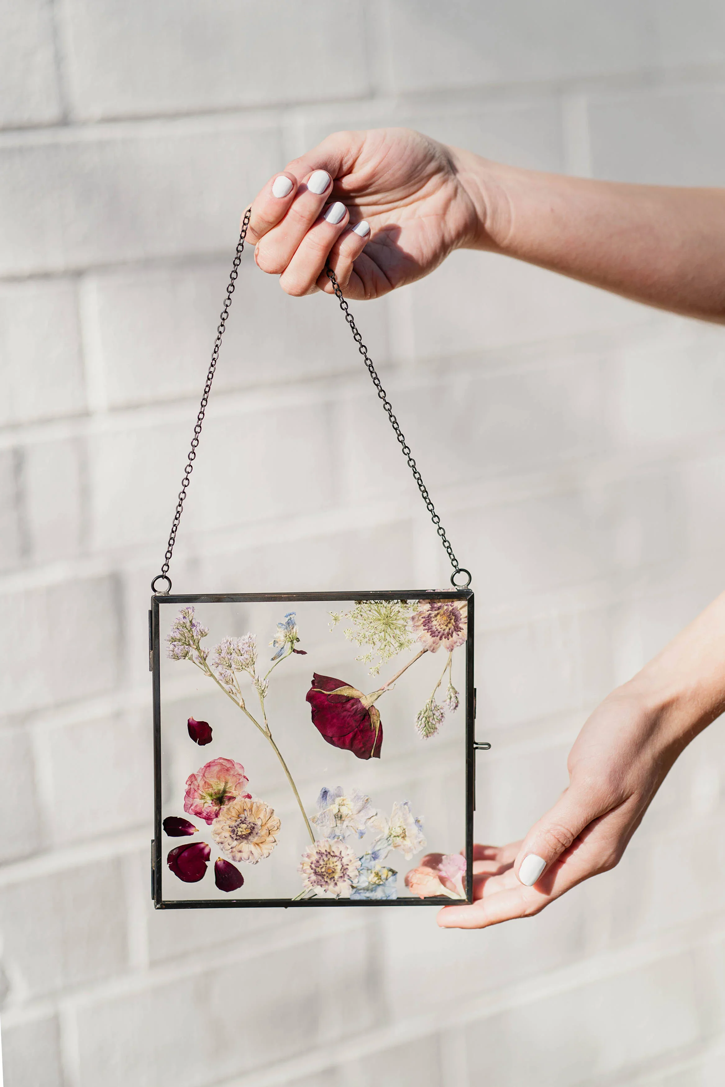A person holding a square glass jewelry case with pressed flowers inside, against a light-colored brick wall.