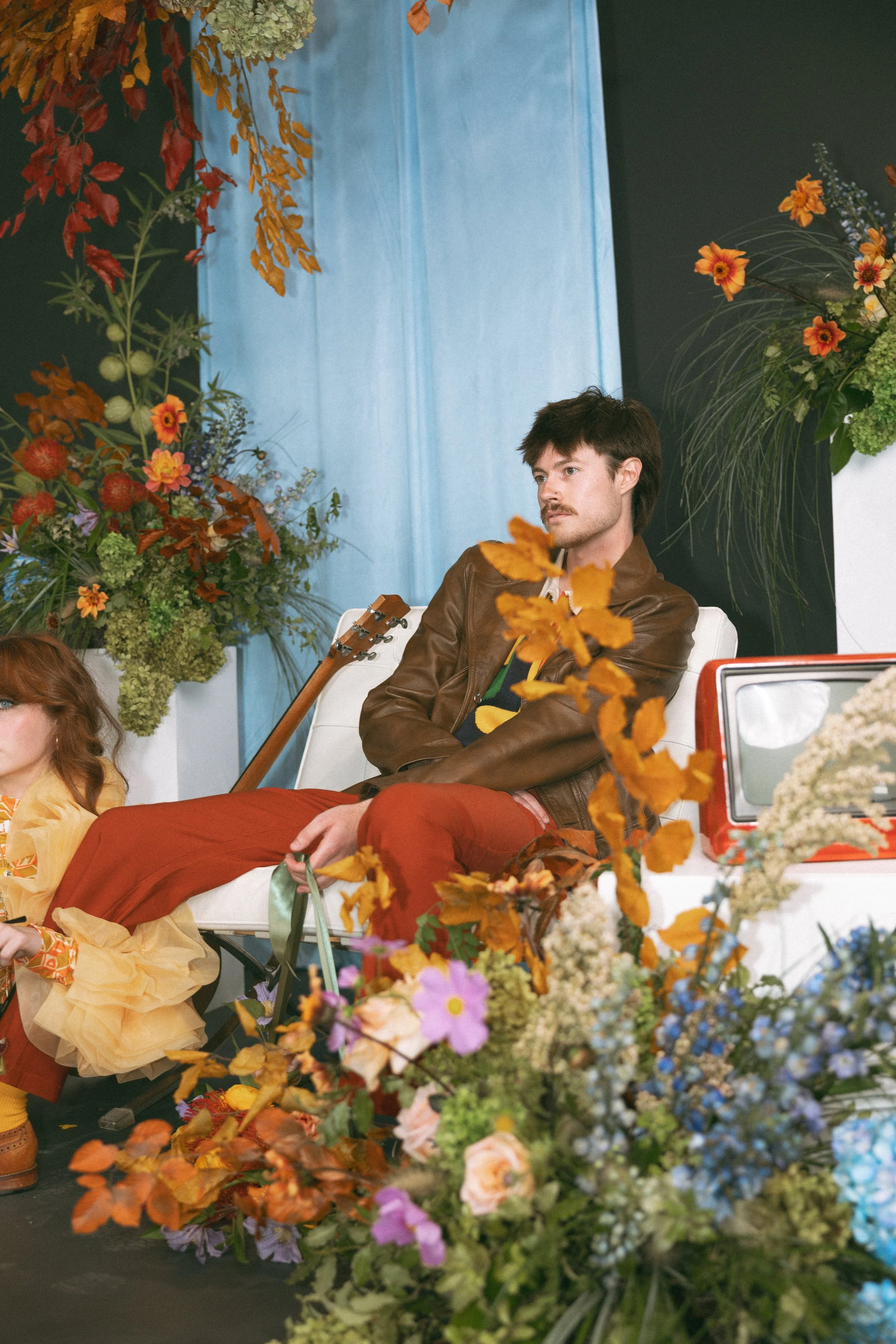 Man sitting on a white bench surrounded by colorful flowers and greenery, with a brown leather jacket and red pants, and a guitar resting behind him.