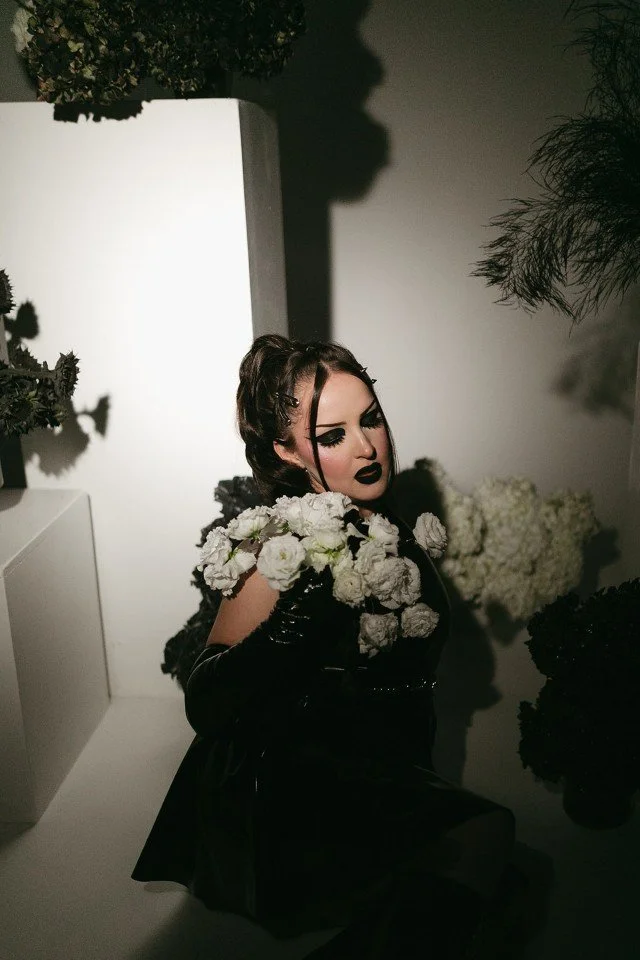 A woman with dark makeup and black lipstick pose in a dimly lit room, surrounded by white flowers and plants, dressed in a black outfit with shiny gloves.
