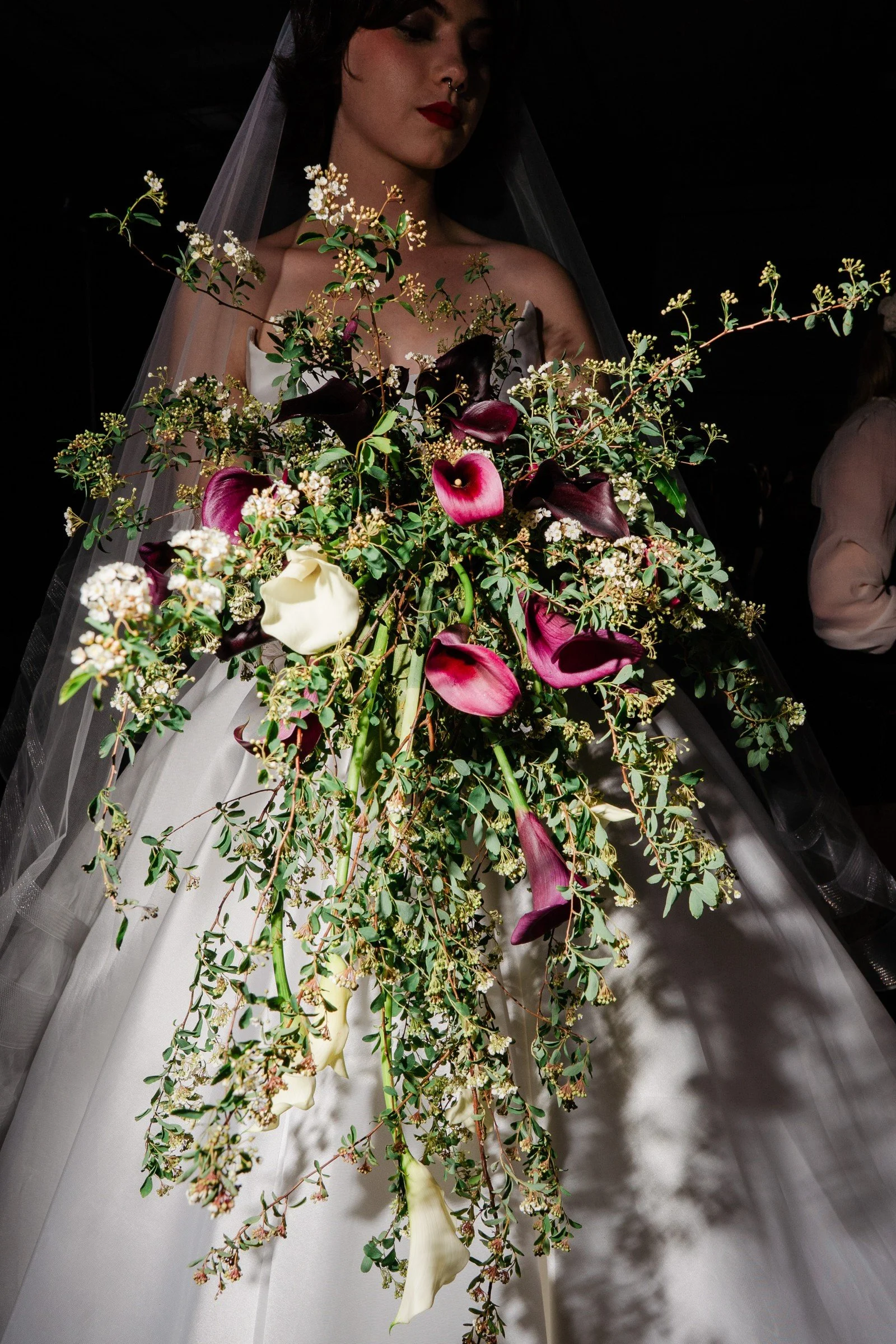 A woman in wedding attire holding a large, cascading bouquet of dark purple, white, and pink flowers with green foliage, partially obscuring her face.