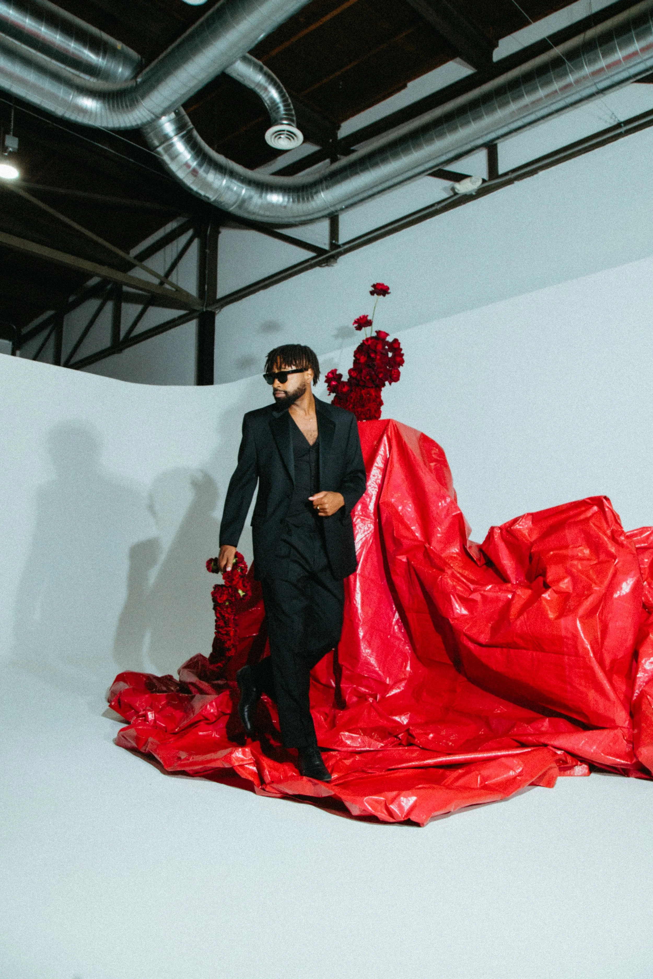 a Black male model in a dark suit against a background of white with a red backdrop and red carnations and roses